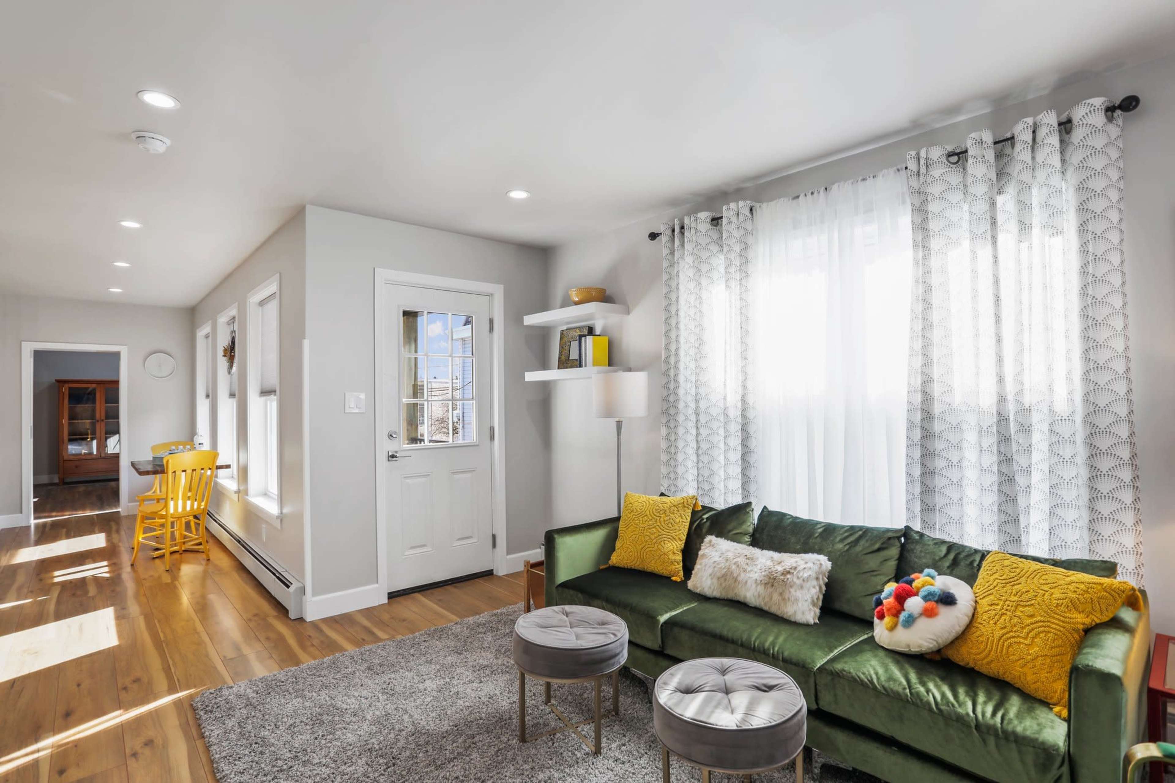 A modern living room with a green sofa, two round silver ottomans, and a bright entrance with large windows and light-colored walls.