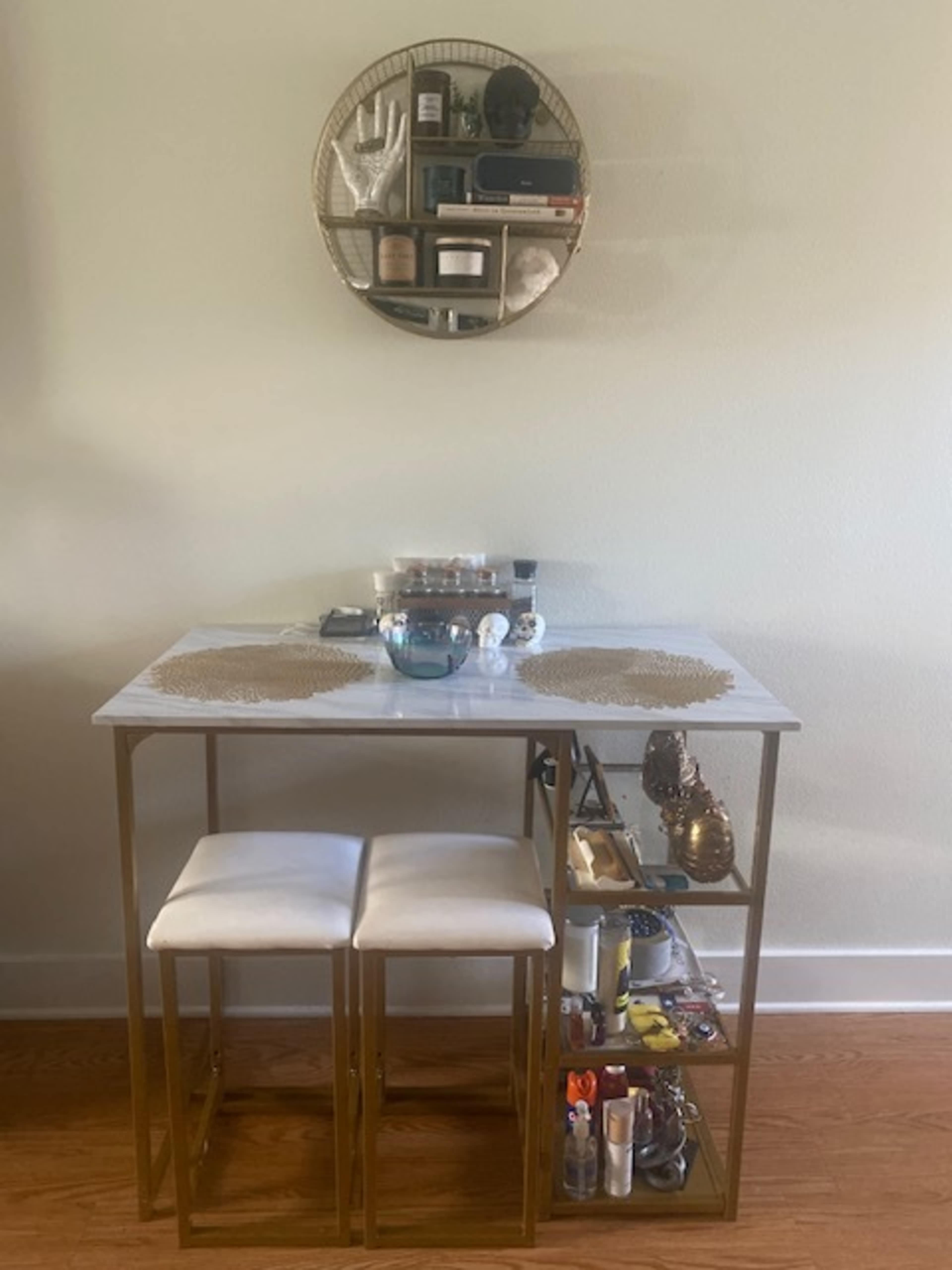 A small dining table with two white stools is placed against a wall, featuring a neatly arranged display of cosmetics and decorative items on shelves above.