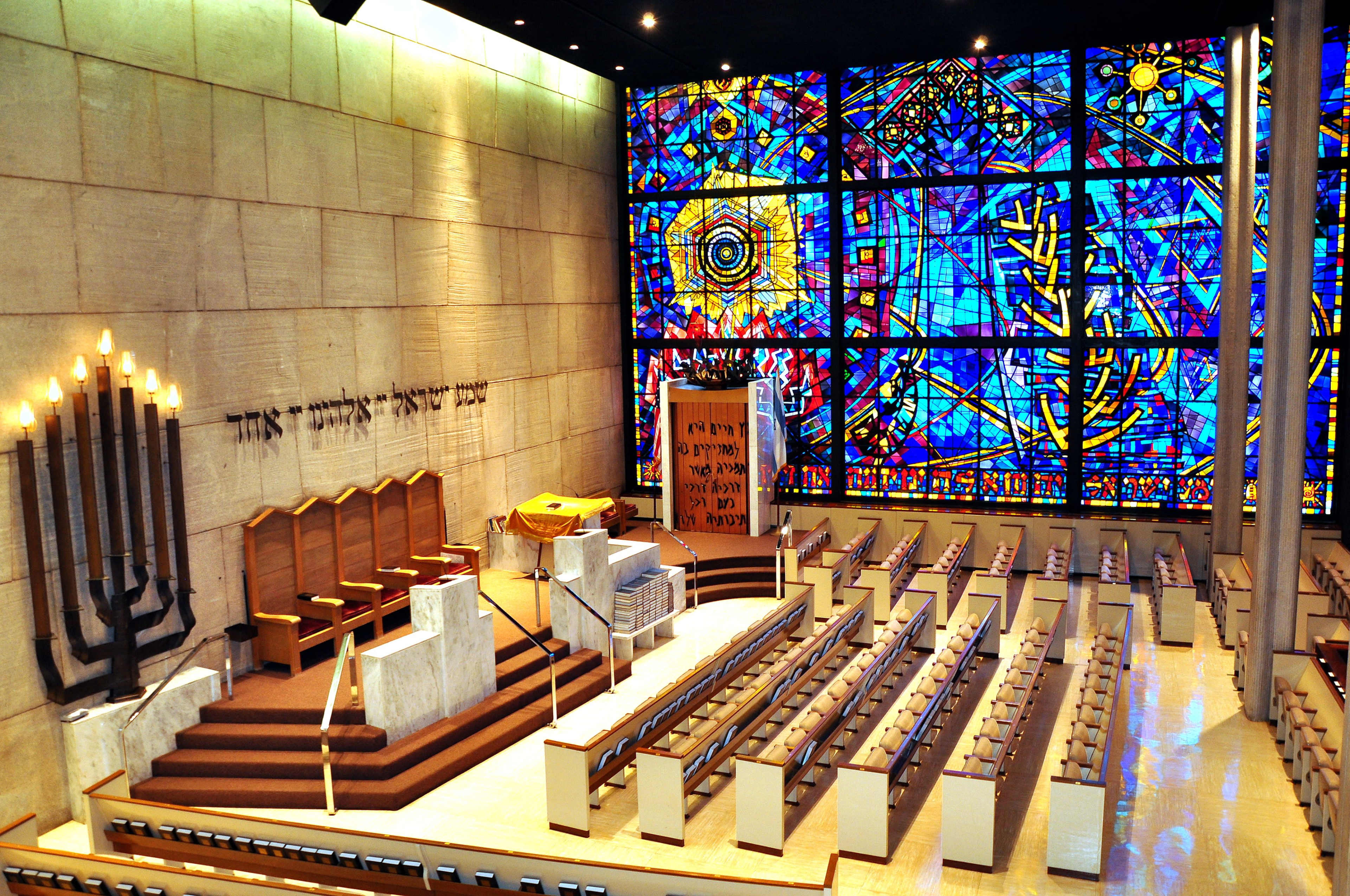 The image shows a spacious synagogue interior featuring a large stained glass window, a raised platform with seating at the front, and rows of wooden benches for worshippers.