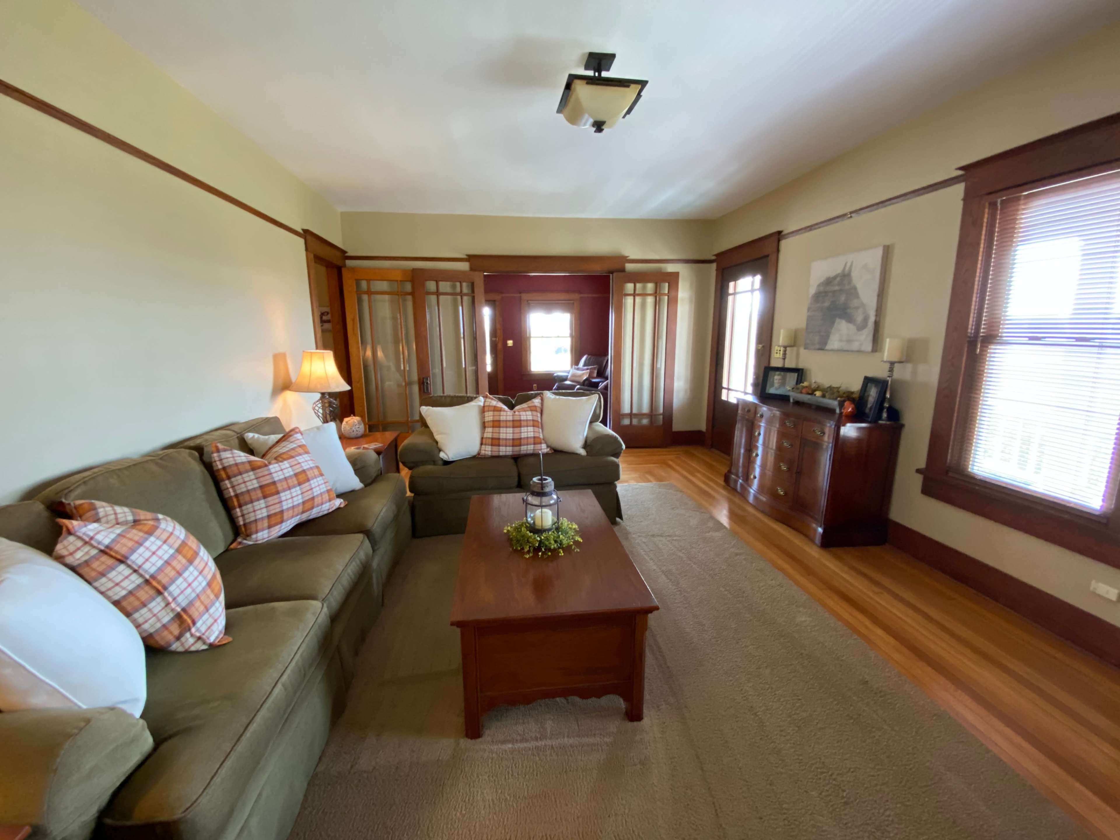 The living room features a green sofa with decorative pillows, a wooden coffee table, and large windows with wooden trim.