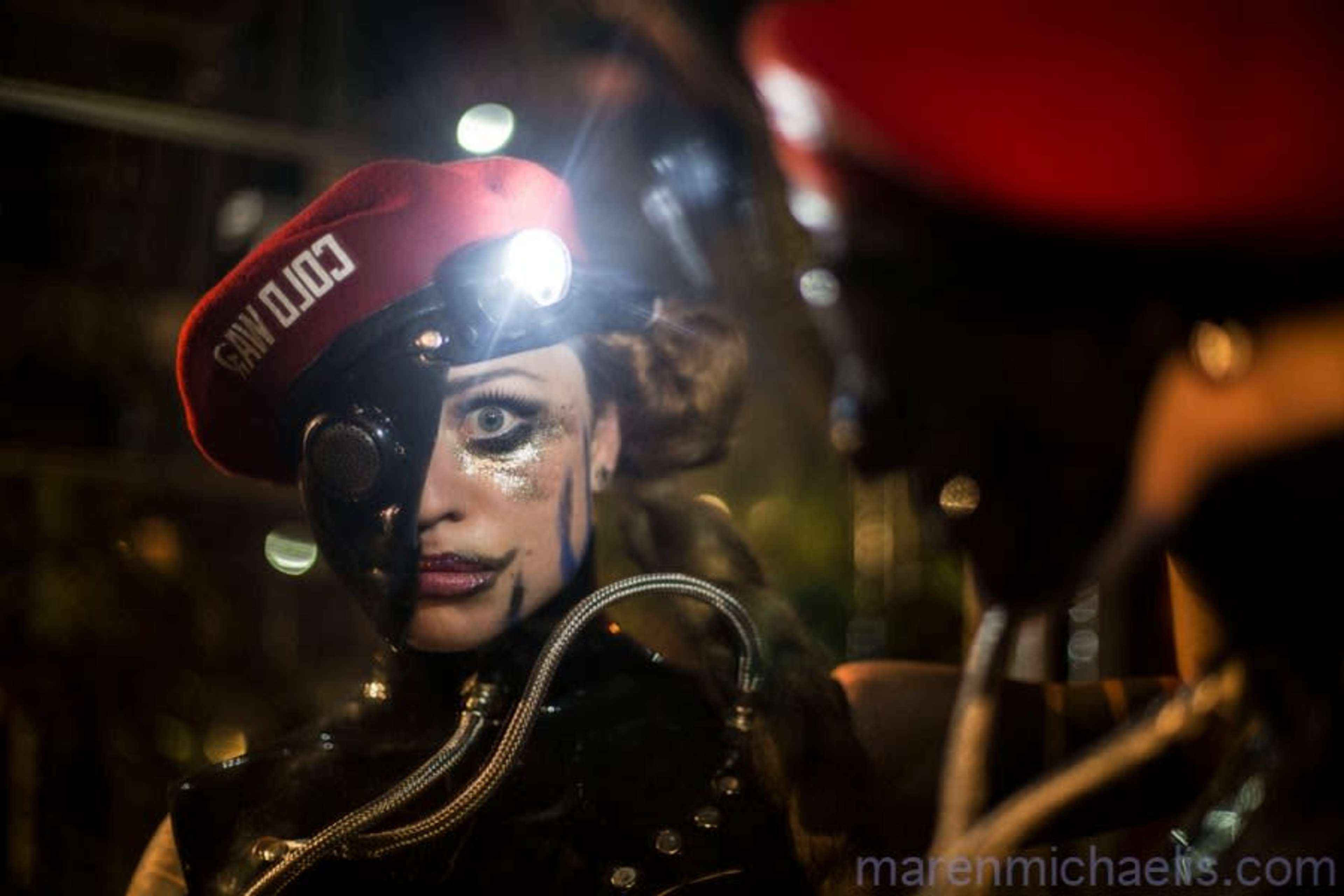 A person wearing a red beret and a metallic eye piece gazes into a reflective surface amid a dimly lit background.