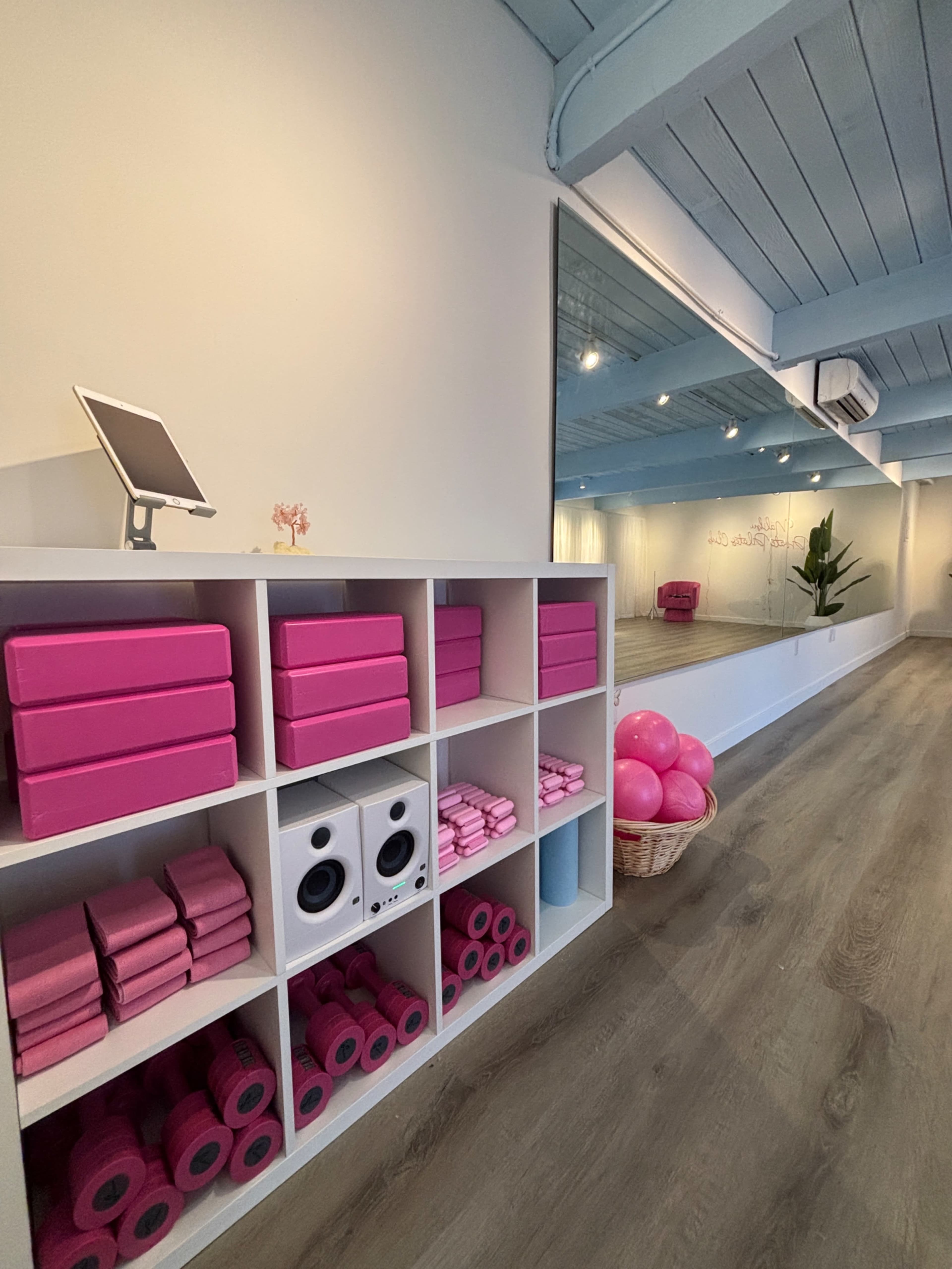 The image shows an interior fitness space featuring pink exercise equipment neatly organized on a white shelf, with a mirrored wall and wooden flooring.