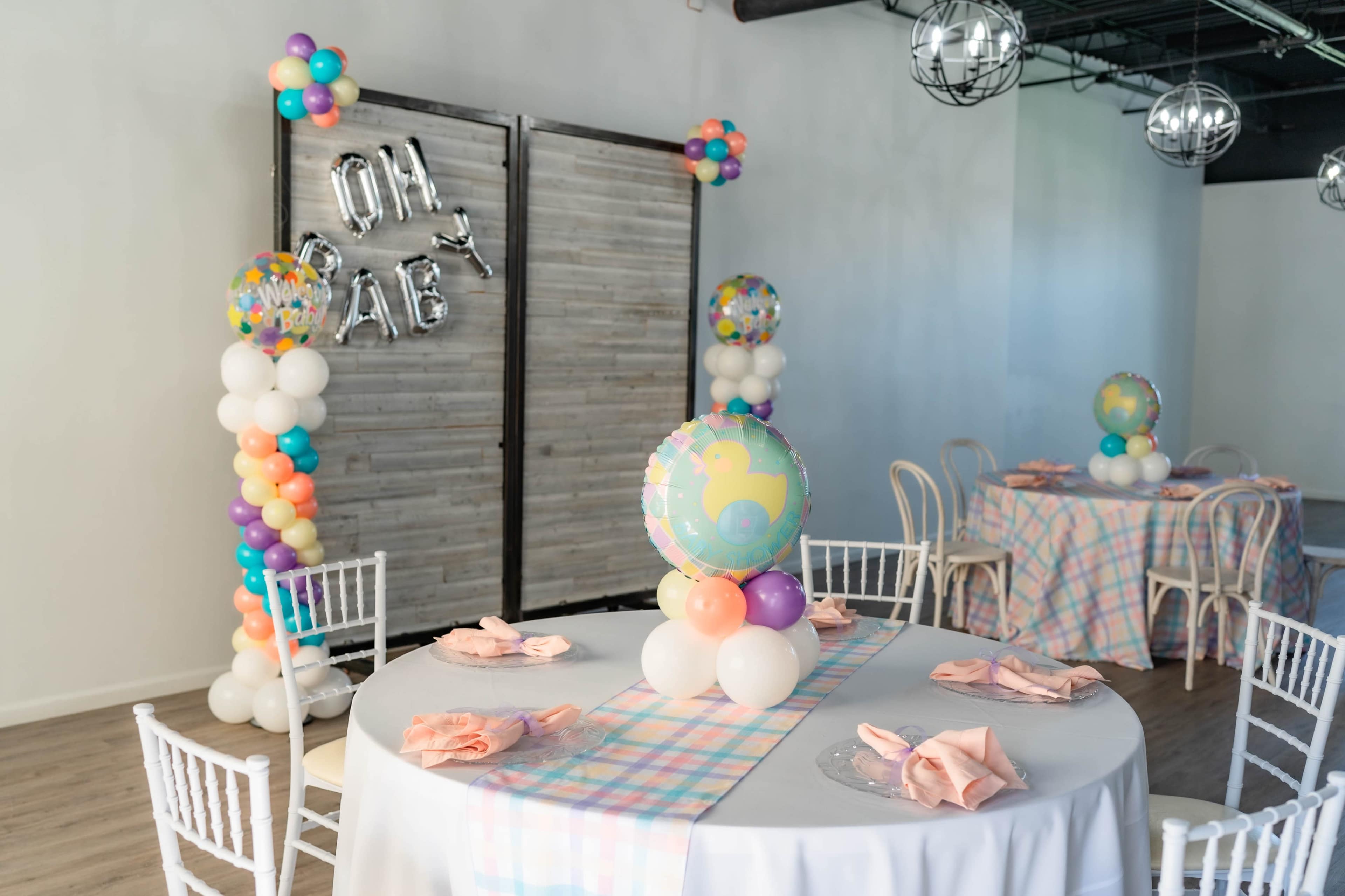 The scene shows a decorated event space with two tables set for a celebration, featuring pastel-colored tablecloths, balloons, and a backdrop with the phrase "Oh Baby."
