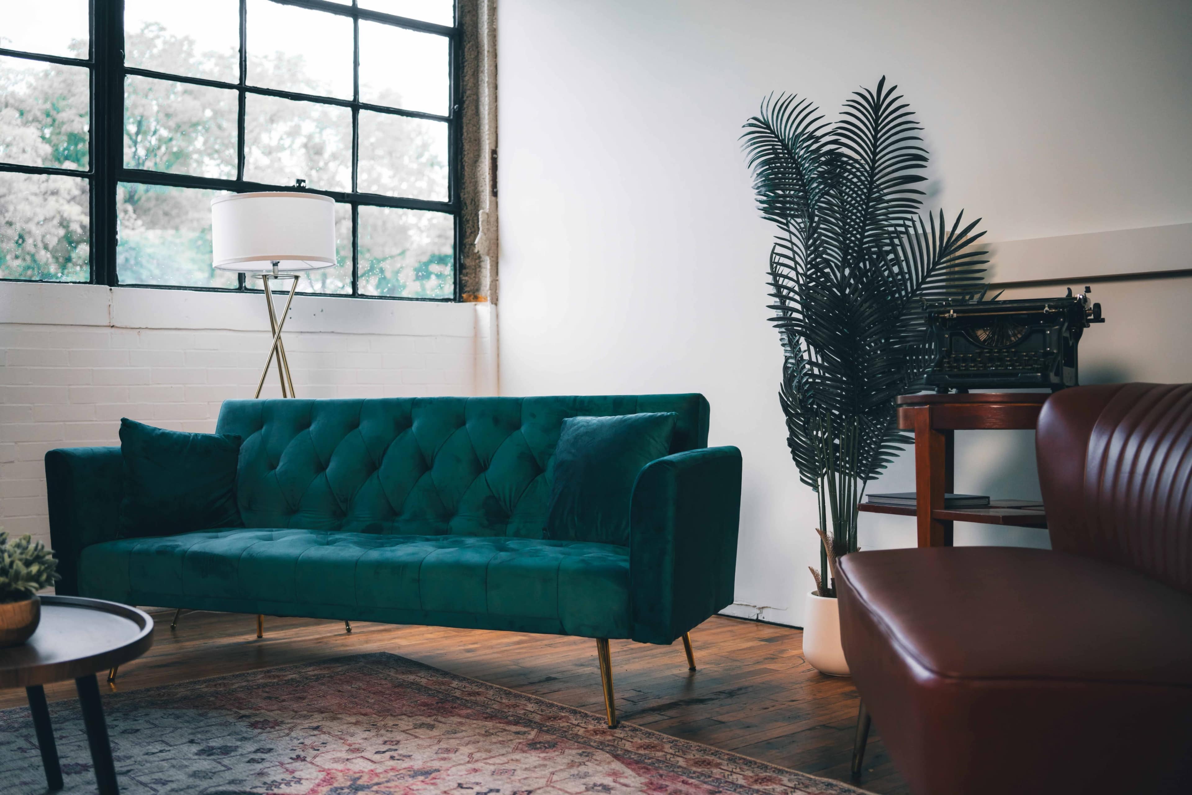 The image shows a modern living room with a green velvet sofa, a tall plant, a floor lamp, and a vintage typewriter on a wooden table.