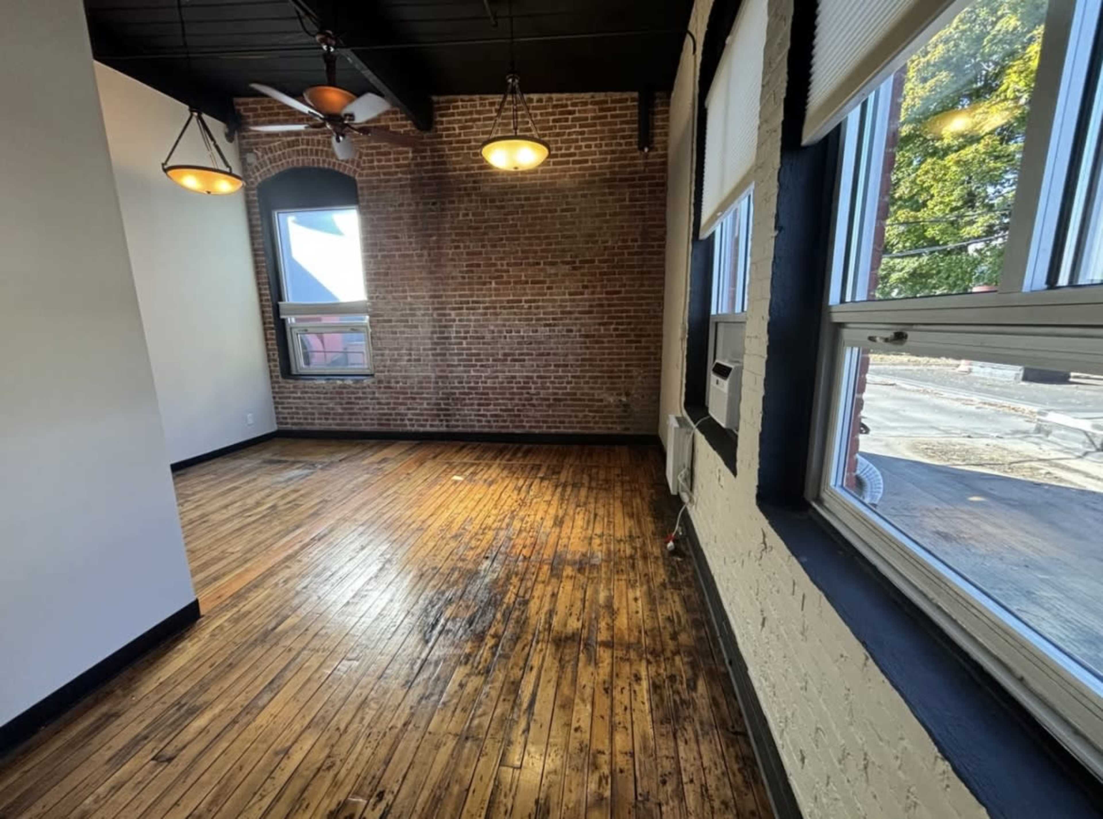 The image shows an empty room with exposed brick walls, large windows, and polished wooden floors.