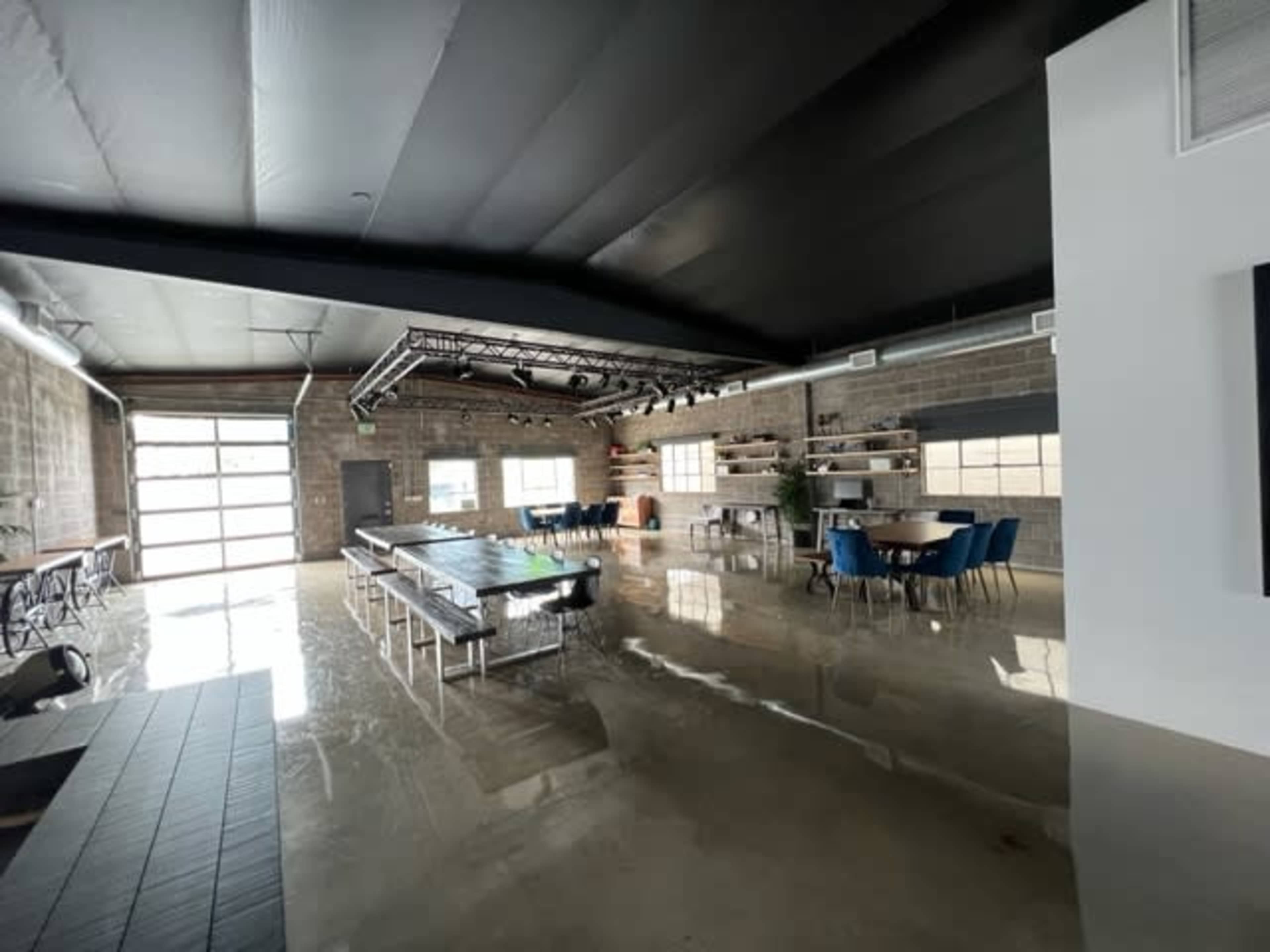 The image shows a spacious, industrial-style room with polished concrete floors, featuring a long central table, several dining tables with blue chairs, and stage lighting overhead.