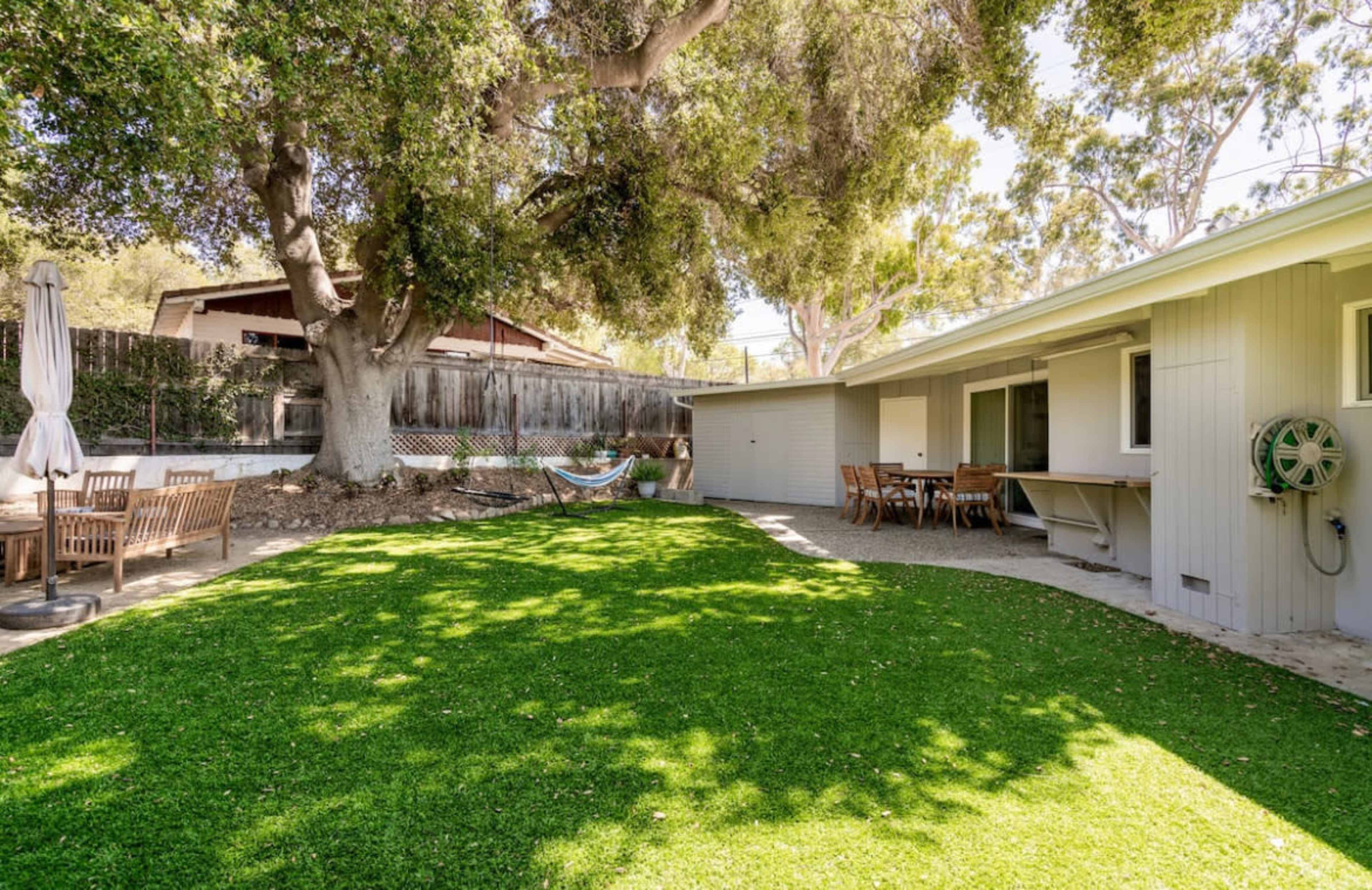 A backyard with a green lawn, a large tree, outdoor seating, and a hammock strung between two posts.