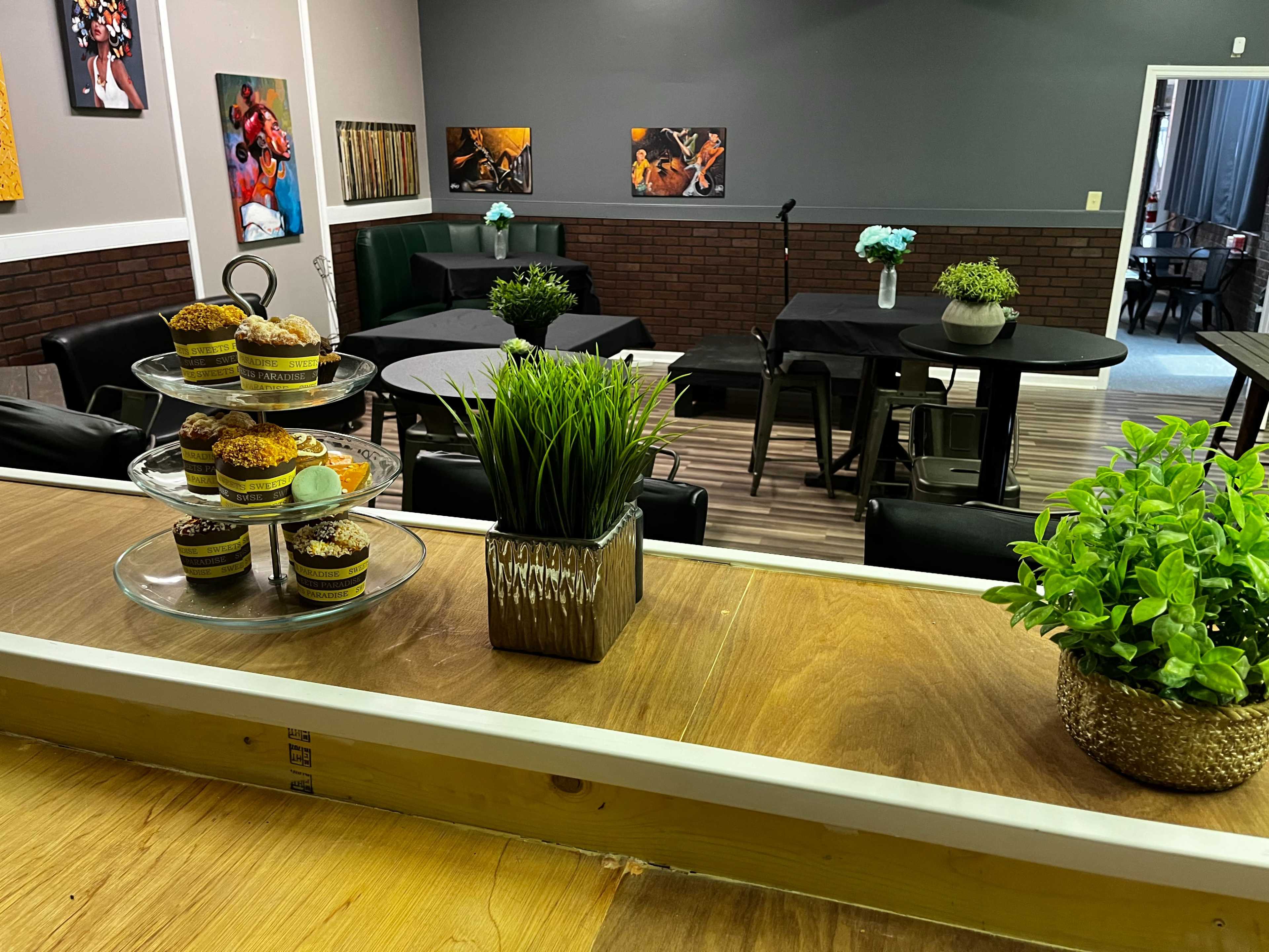 The image shows a stylized room with tables and chairs arranged for an event, featuring a display of cupcakes on a stand in the foreground and potted plants on the table.