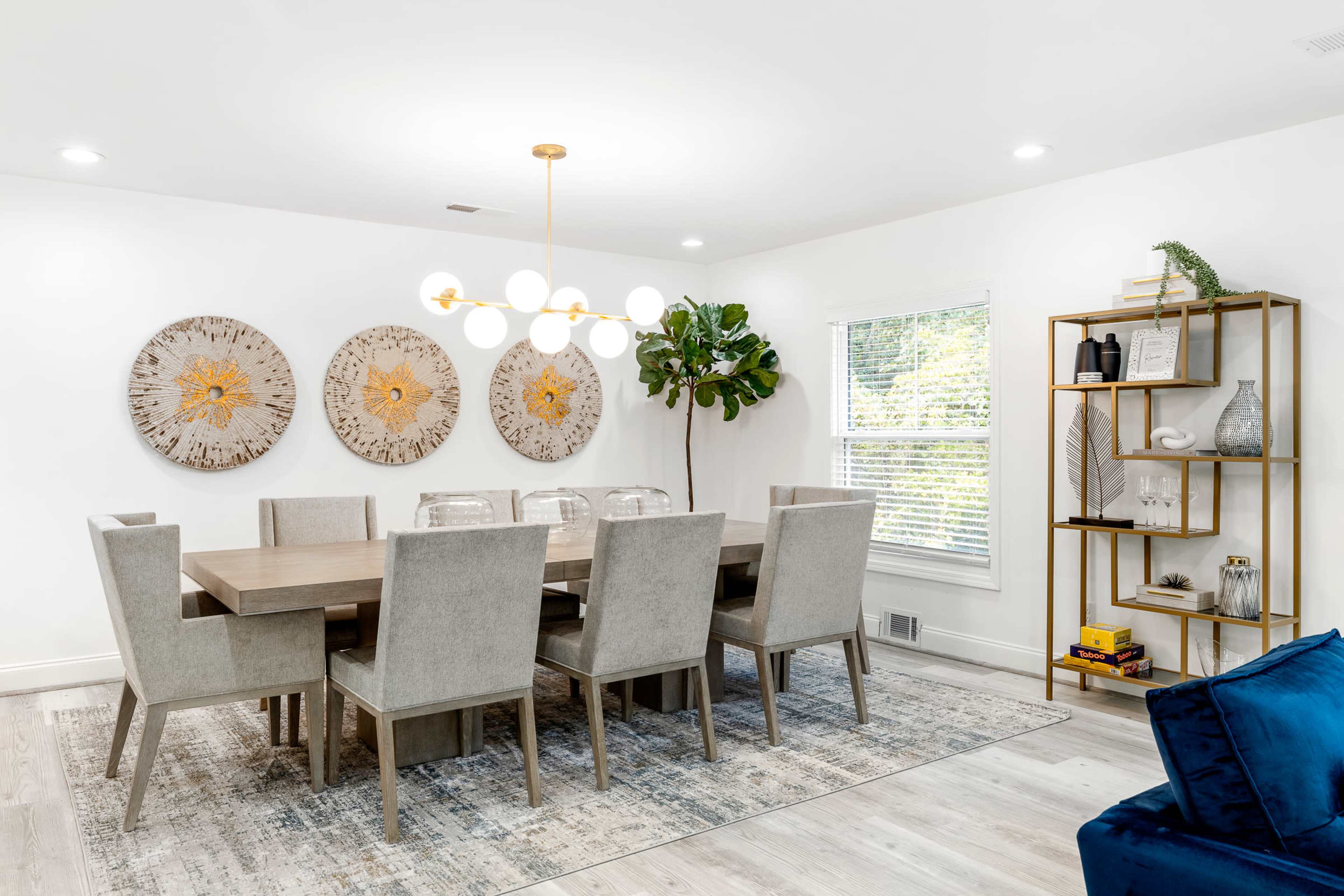 A modern dining room features a long wooden table surrounded by upholstered chairs, with three decorative wall hangings and a bookshelf against the wall.