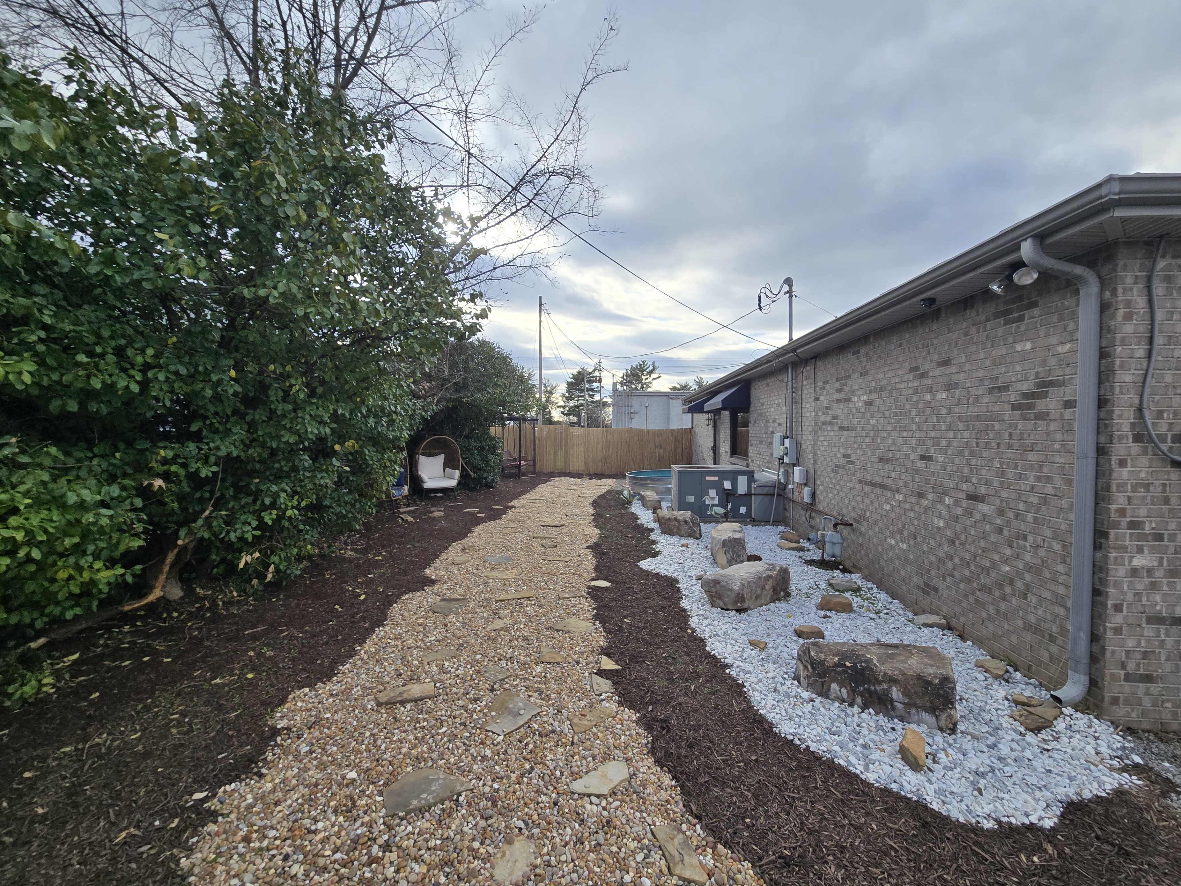 The image shows a pathway leading through a landscaped yard, lined with gravel and mulch, with shrubs on one side and a building on the other.