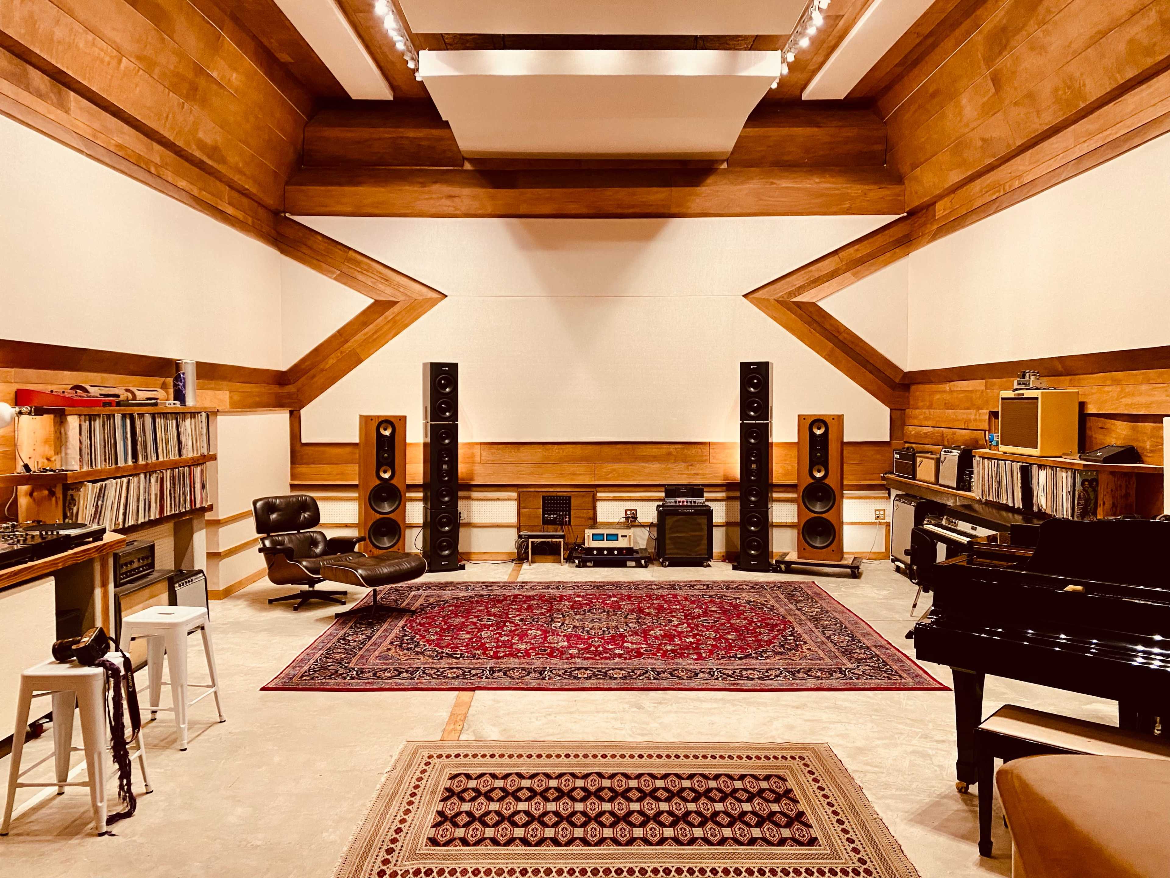 A spacious music studio features vinyl records on shelves, large speakers on either side, a grand piano, and a patterned rug on the floor.