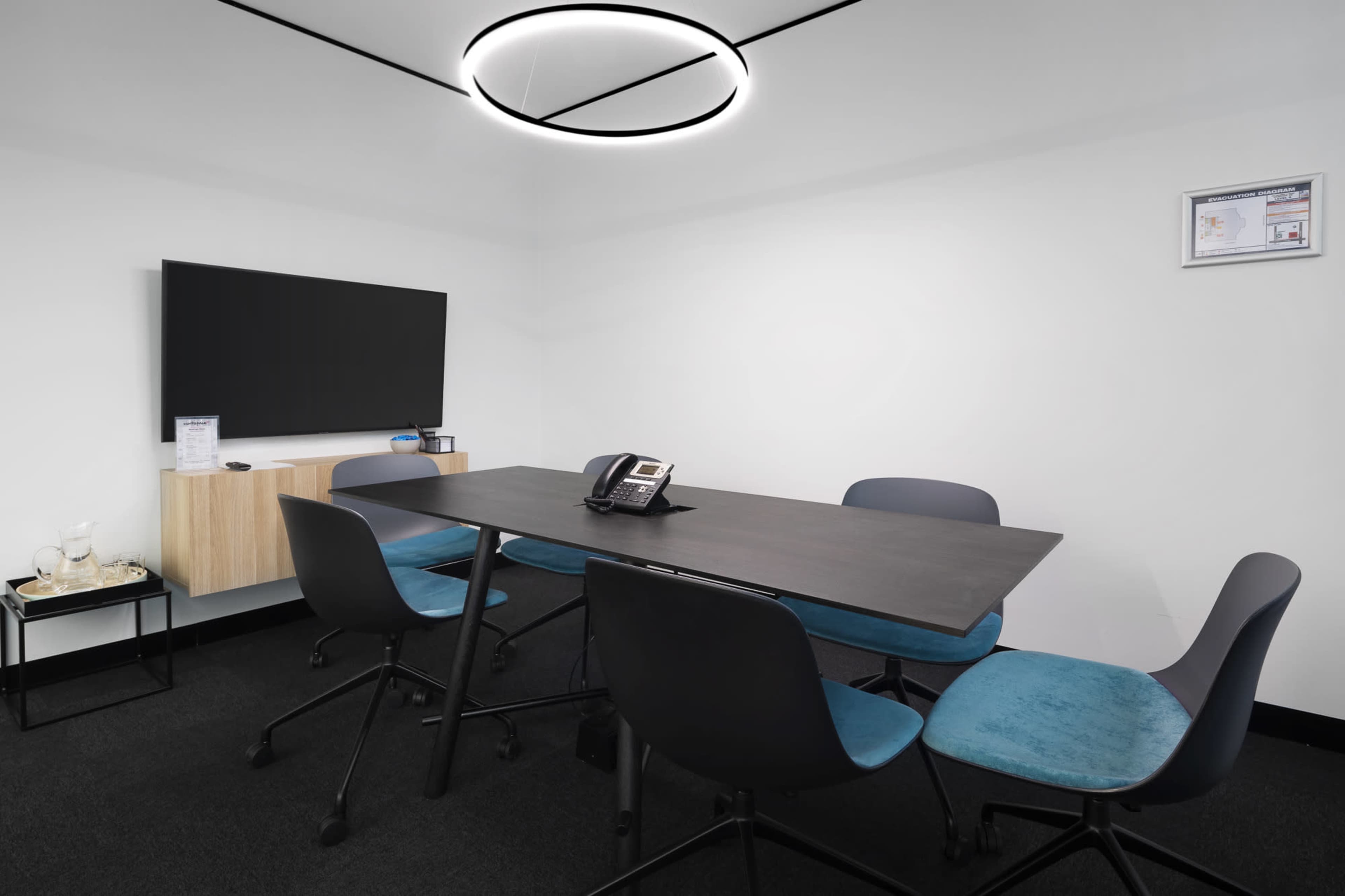 A conference room features a rectangular table surrounded by chairs, a wall-mounted TV, and a modern, circular light fixture.