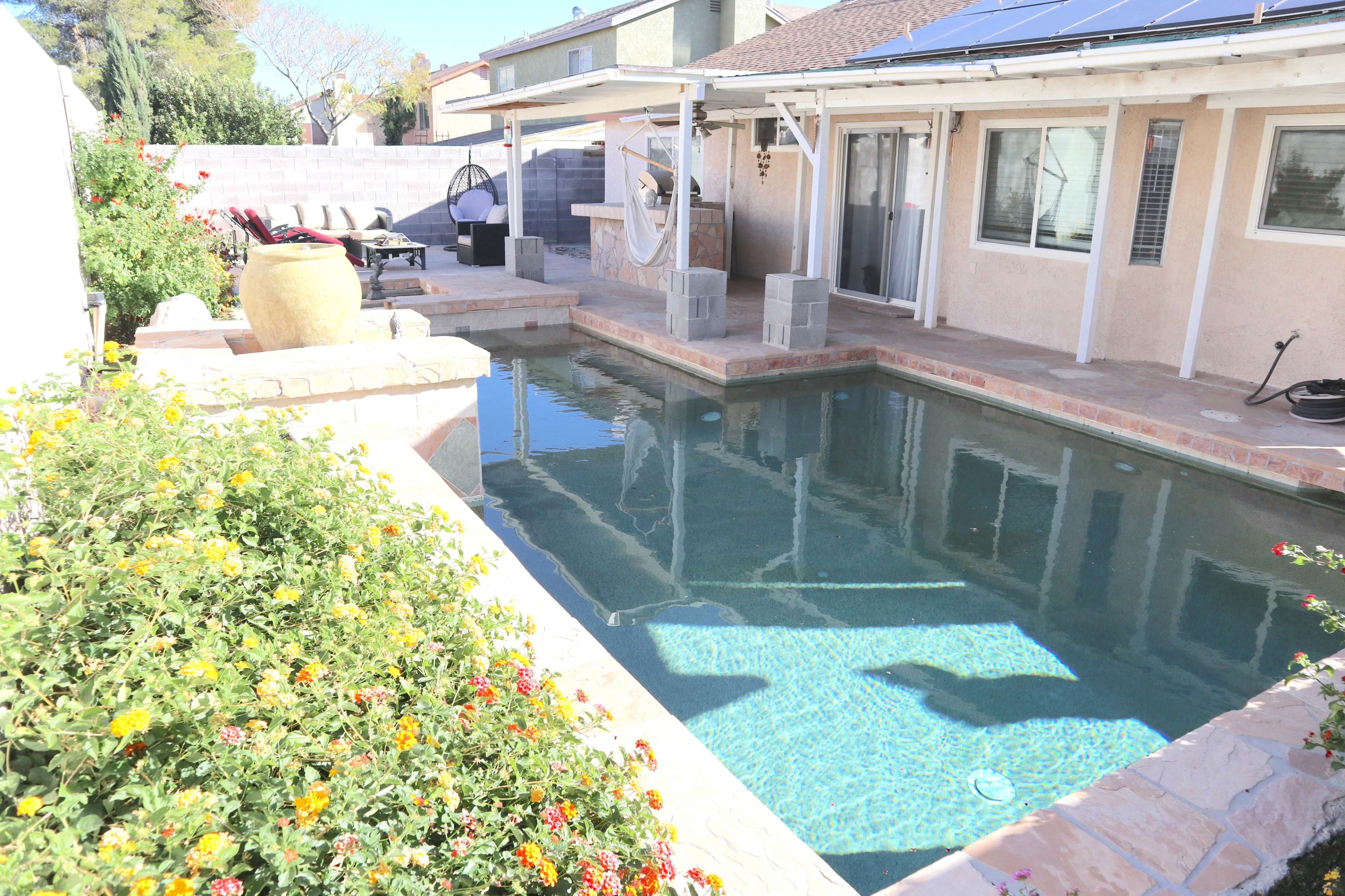 A backyard pool with surrounding flower beds, a patio area with furniture, and a house featuring a solar panel roof.