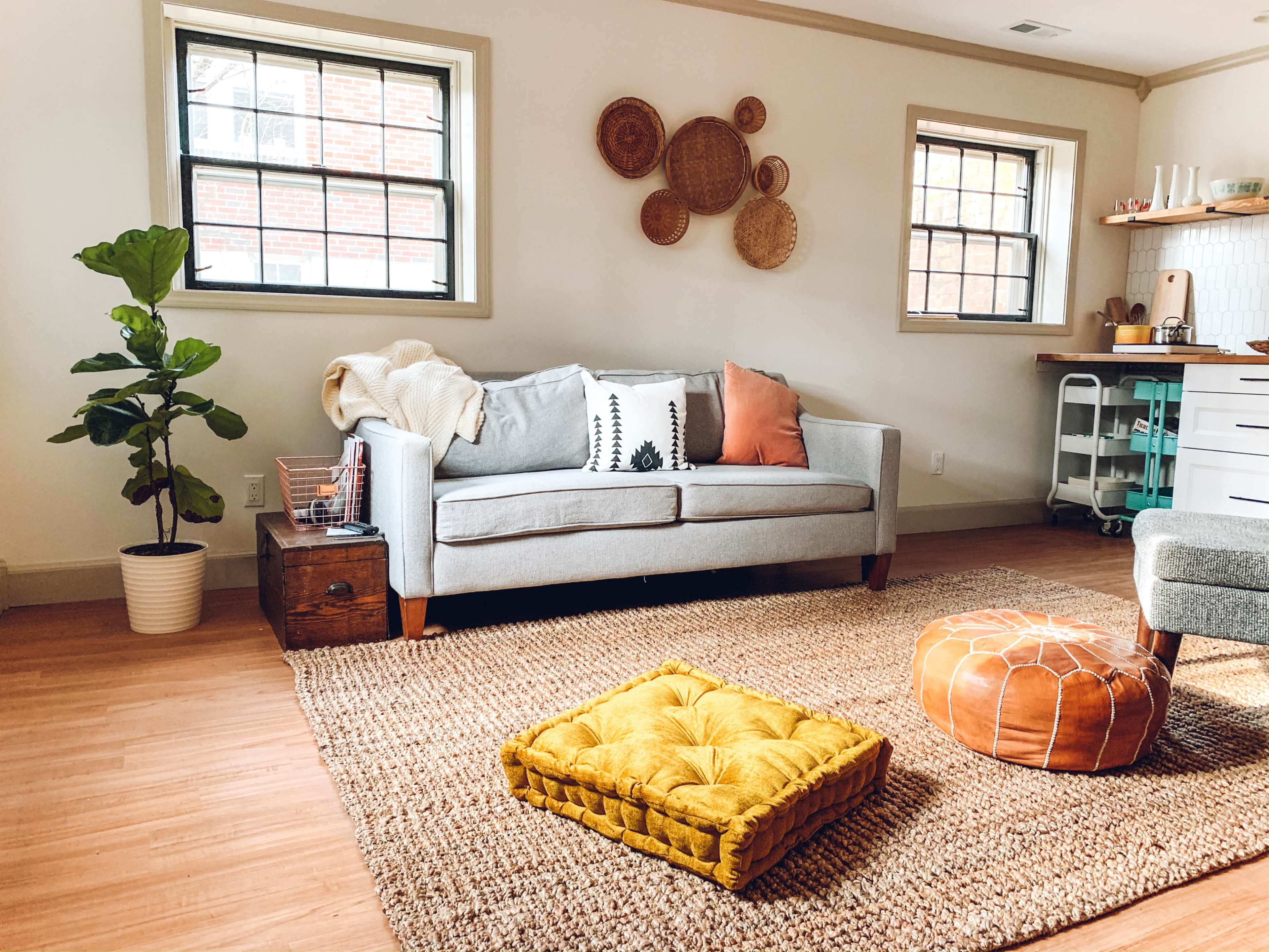 The scene features a cozy living room with a gray sofa, colorful cushions, a round pouf, and decorative wall hangings, set on a woven rug with a potted plant nearby.