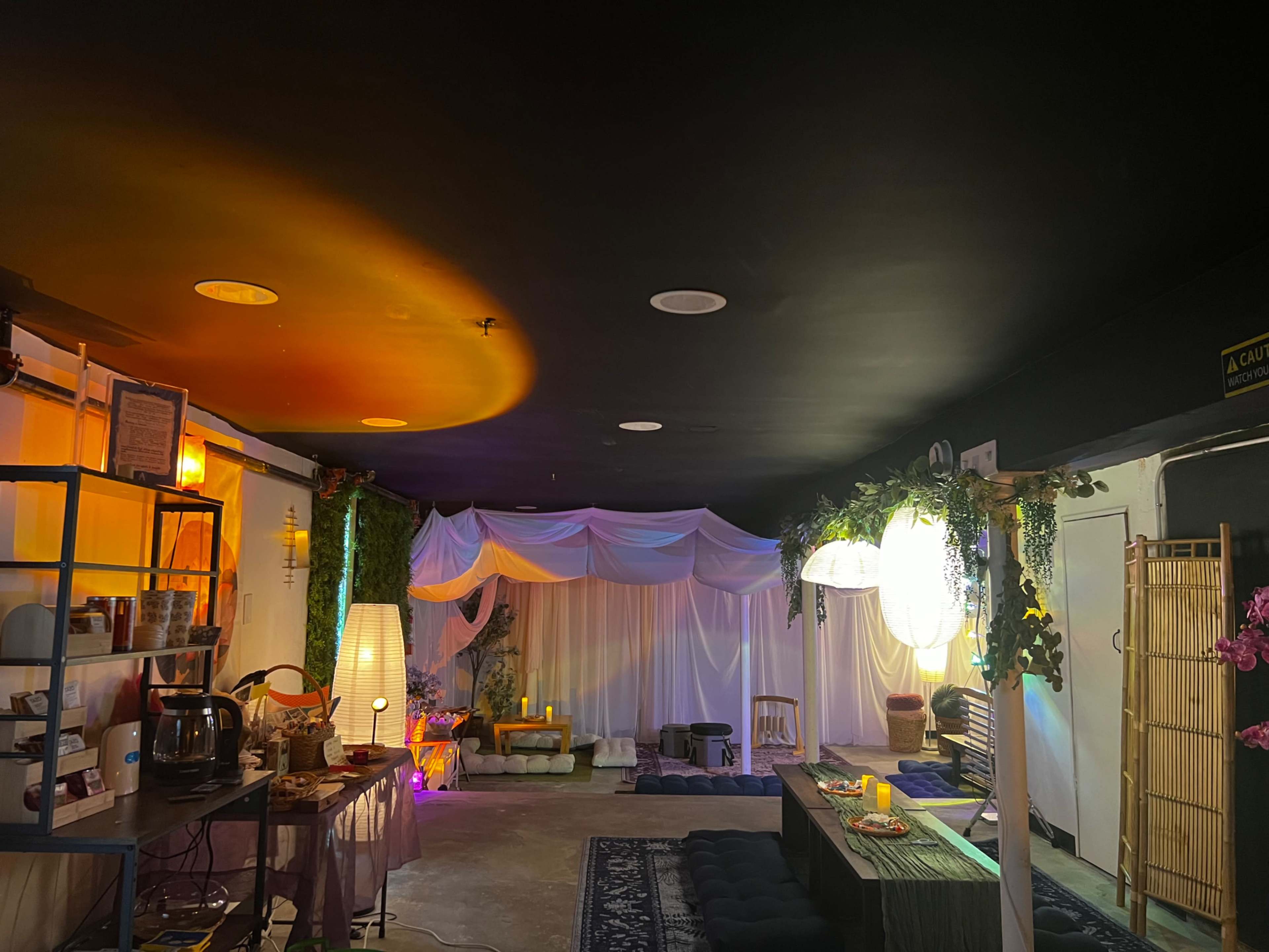 The image shows a cozy interior space with soft lighting, draped fabric overhead, and various seating areas arranged for relaxation.