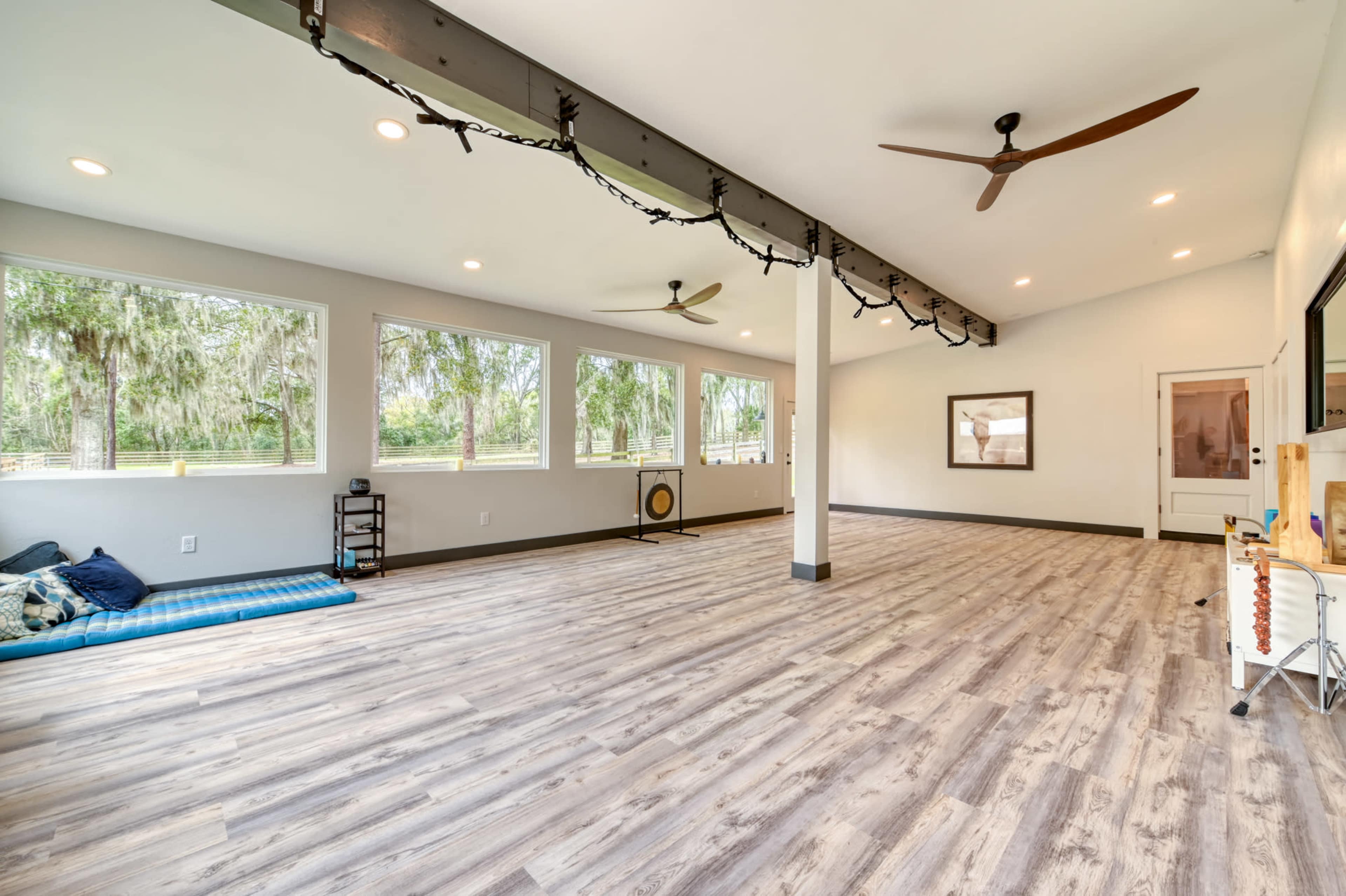 The image shows a spacious, empty room with large windows, wood flooring, and ceiling fans.
