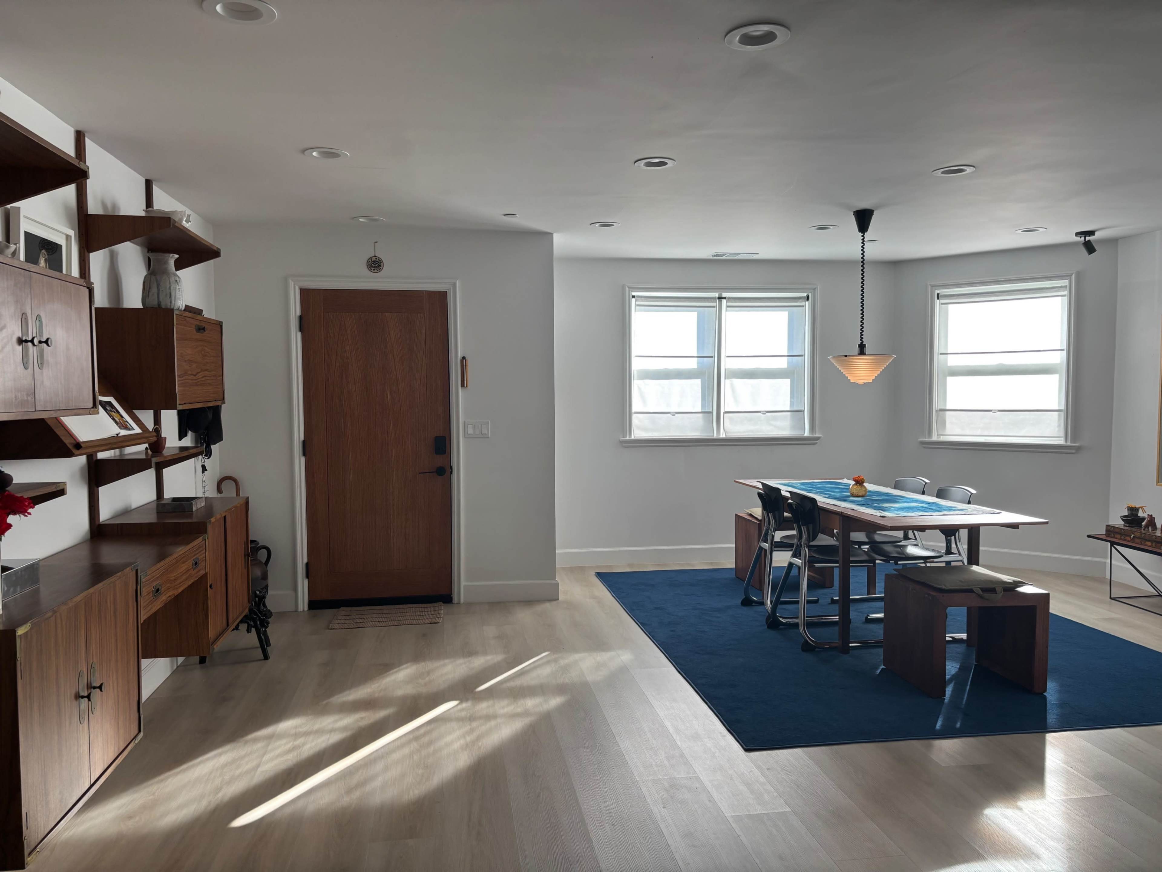 The image shows a modern interior space with a dining area featuring a table and chairs, a blue rug, and a wooden cabinet along the wall.