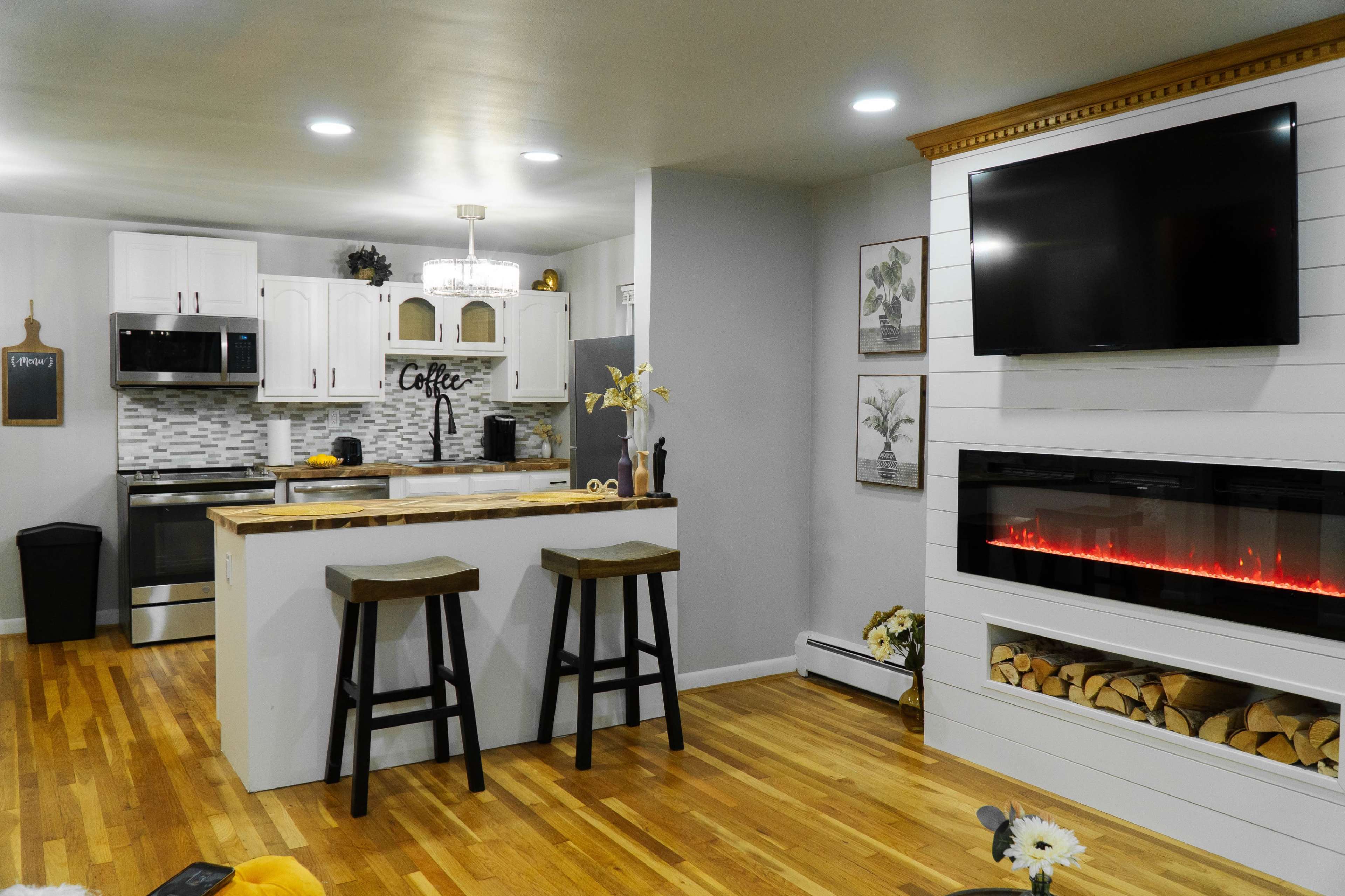 A modern kitchen and living area featuring white cabinetry, a wooden island with two stools, and an electric fireplace with a wood stack beneath a mounted TV.