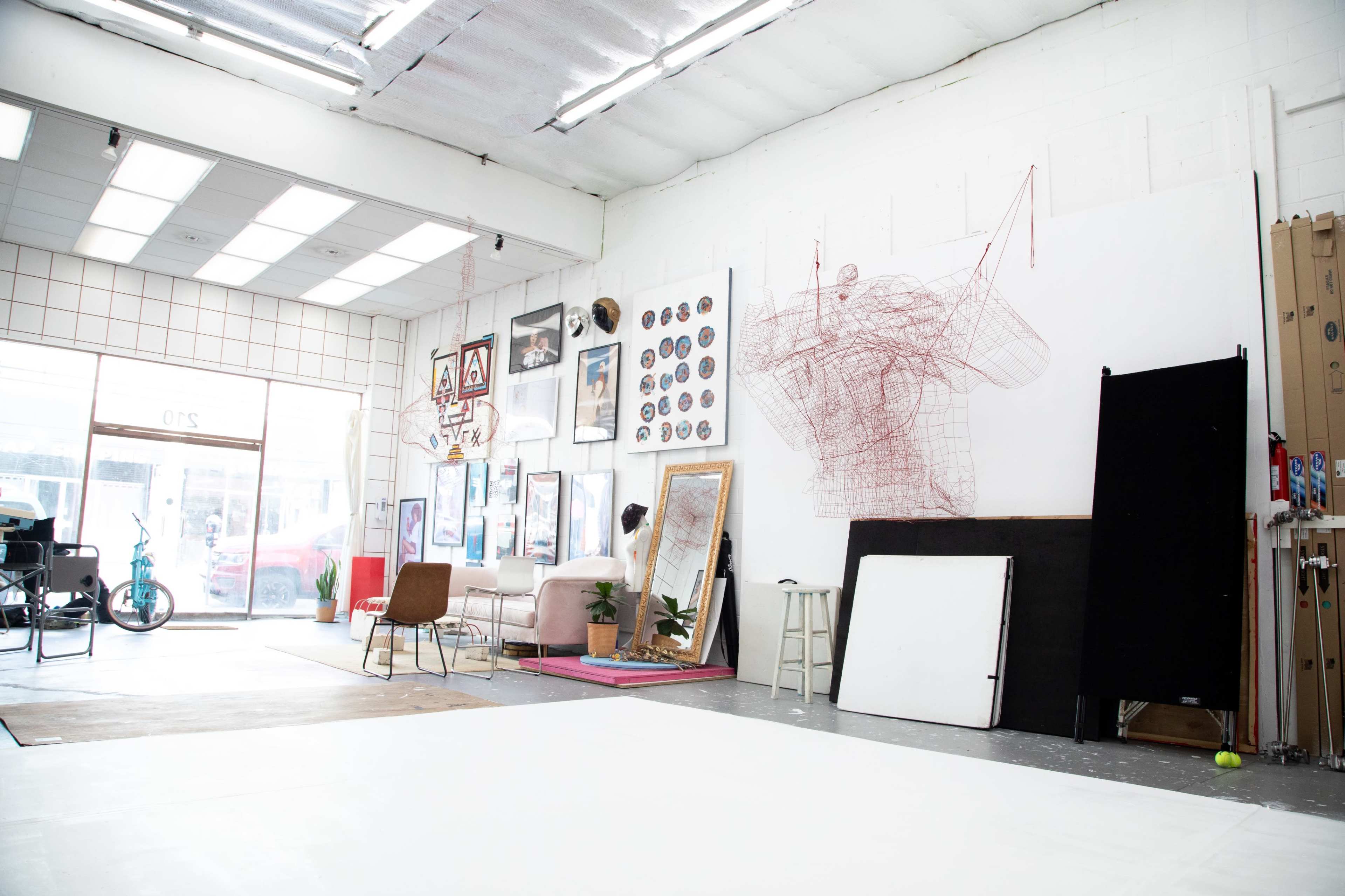 An art studio with large windows, various artworks on the walls, a chair, and easels set up for creative work.