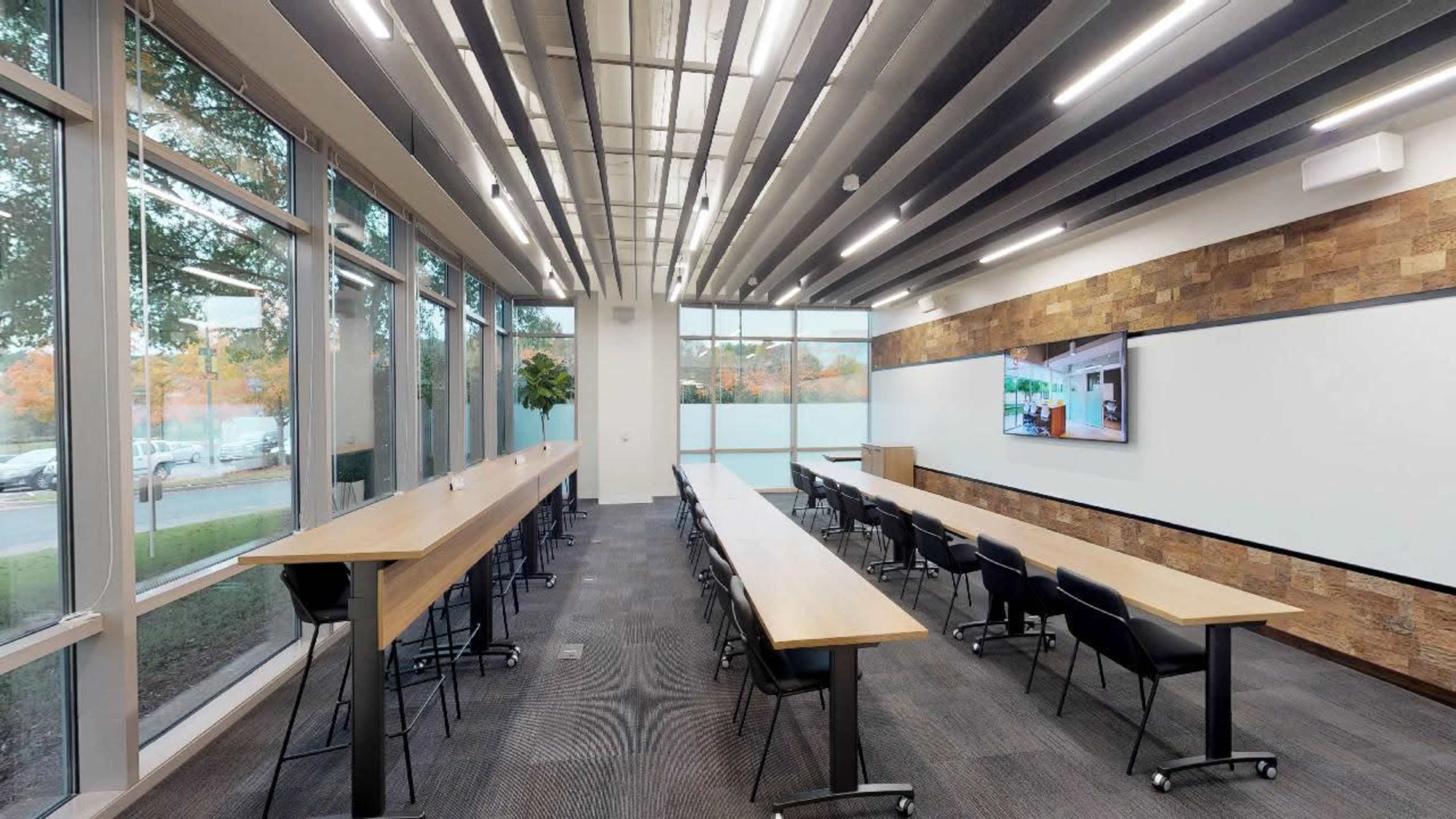 A modern conference room with long tables, chairs, large windows, and a screen displaying digital content.