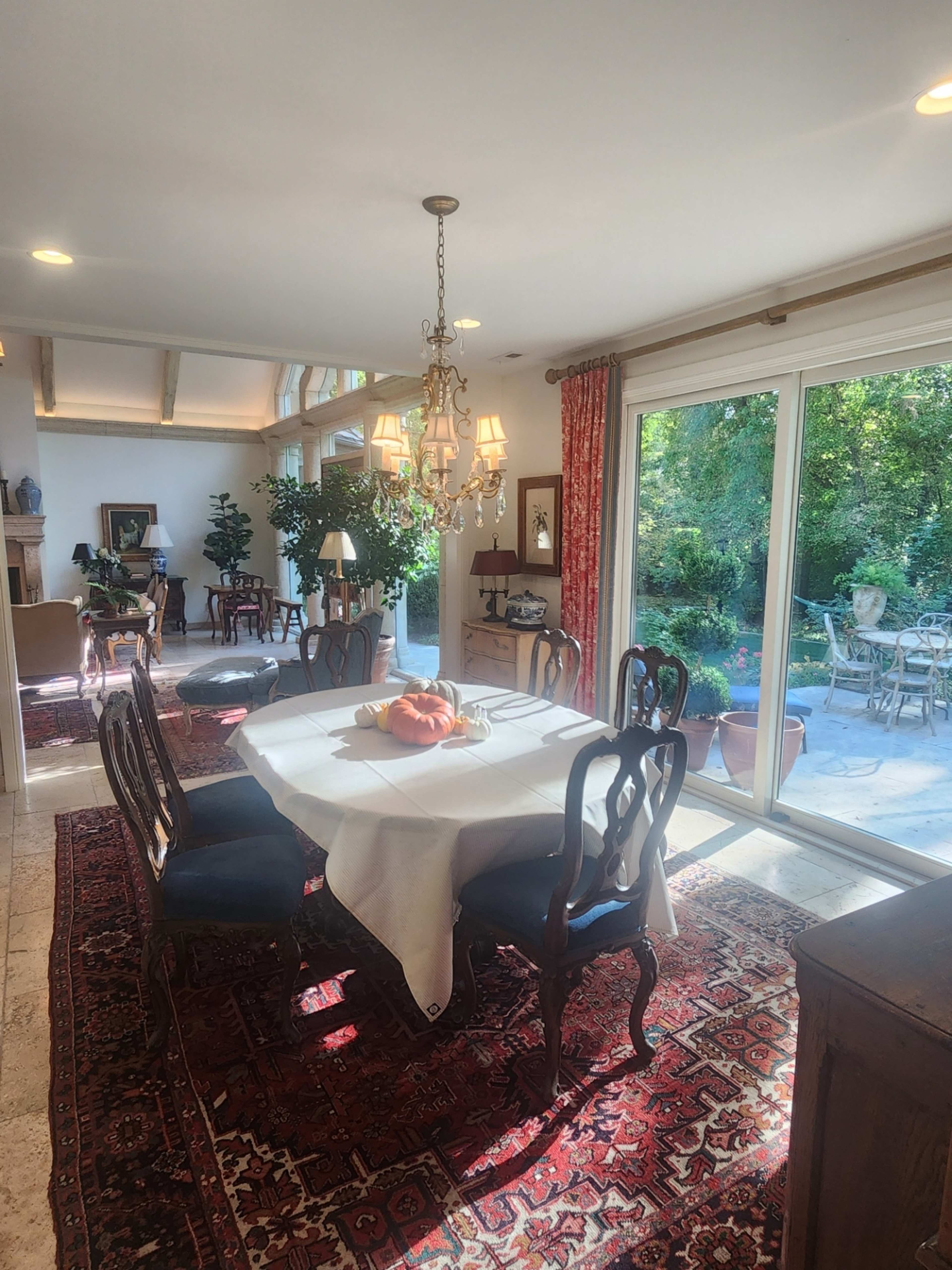 A dining area features a round table with a white tablecloth, surrounded by wooden chairs, and large windows that overlook a garden.