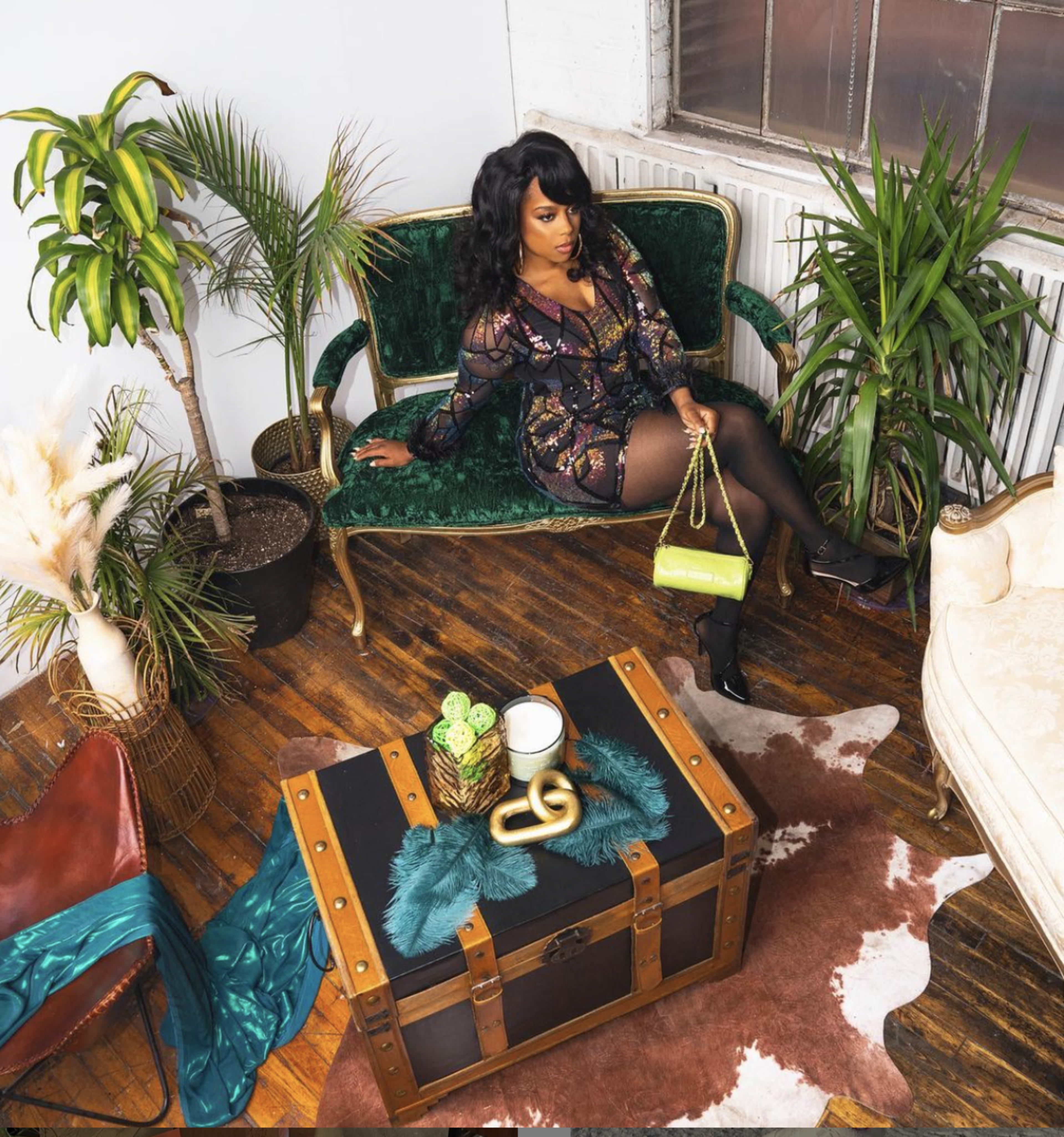 A woman in a patterned dress sits on a green velvet sofa surrounded by potted plants and a decorative coffee table in a well-lit, stylish room.