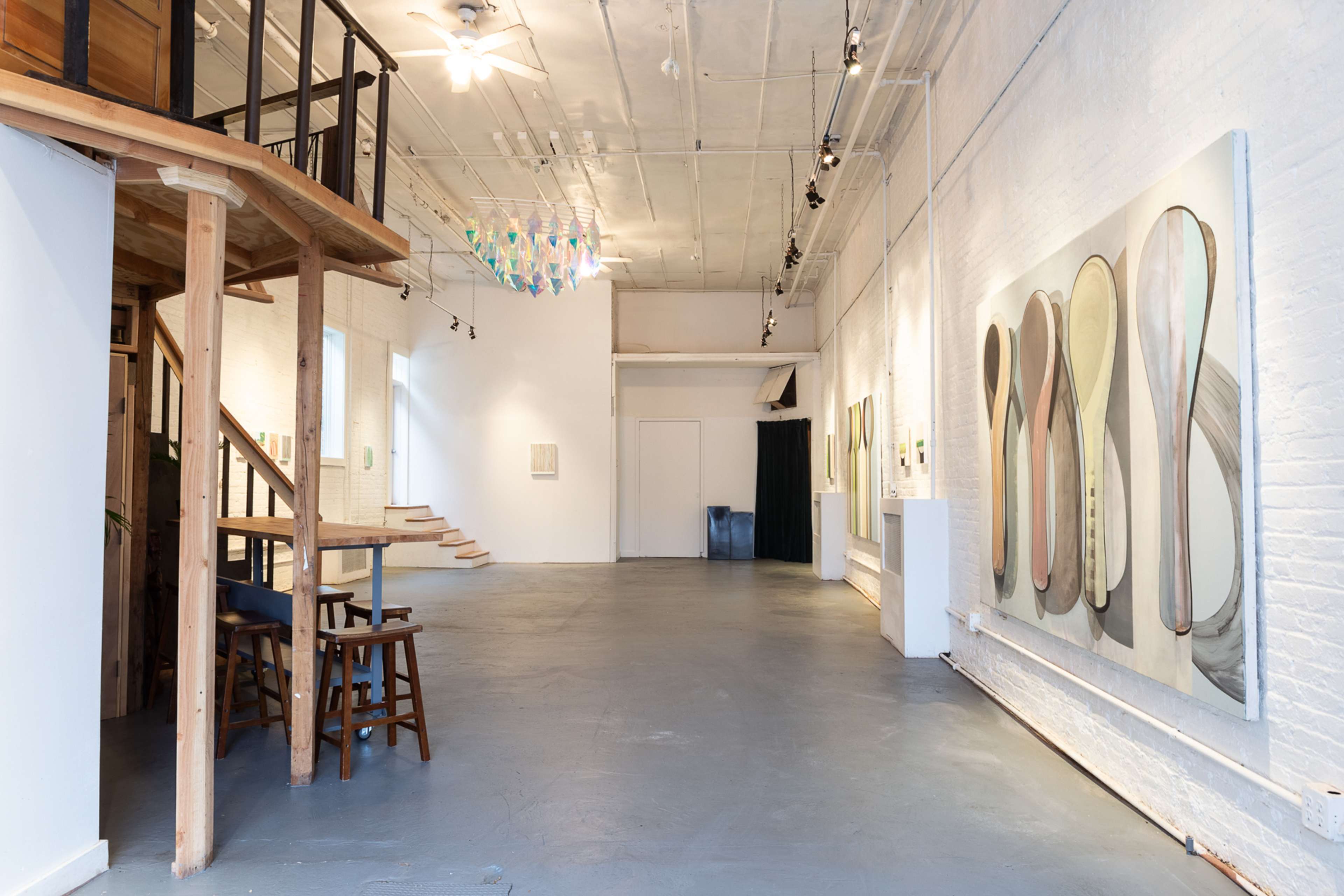 A spacious art gallery with wooden stairs on the left, various artworks on the walls, and colorful hanging sculptures above.