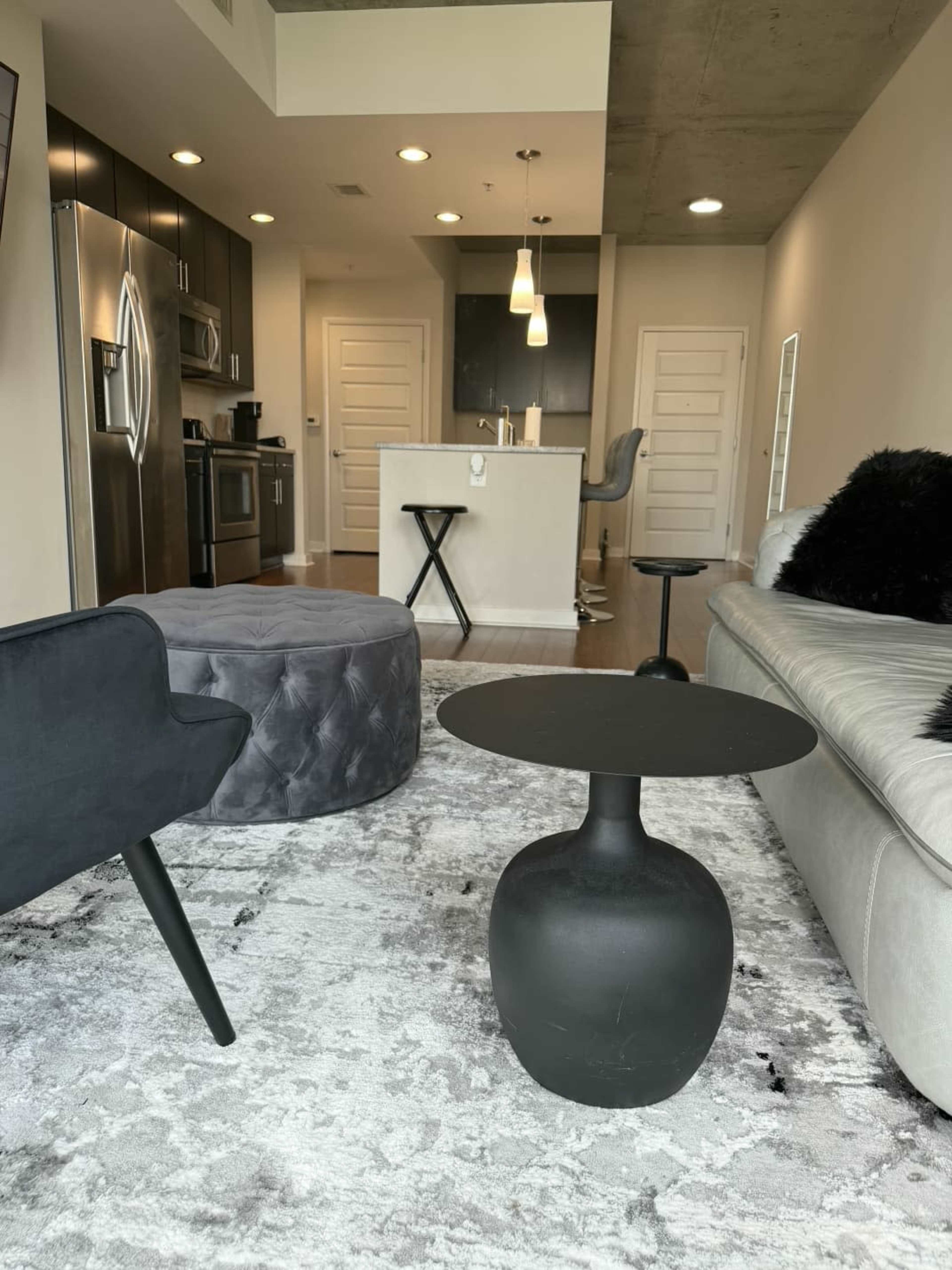 The image shows a modern living area featuring a gray sofa, a round black coffee table, two armchairs, and a plush gray ottoman, all situated on a light-colored area rug.