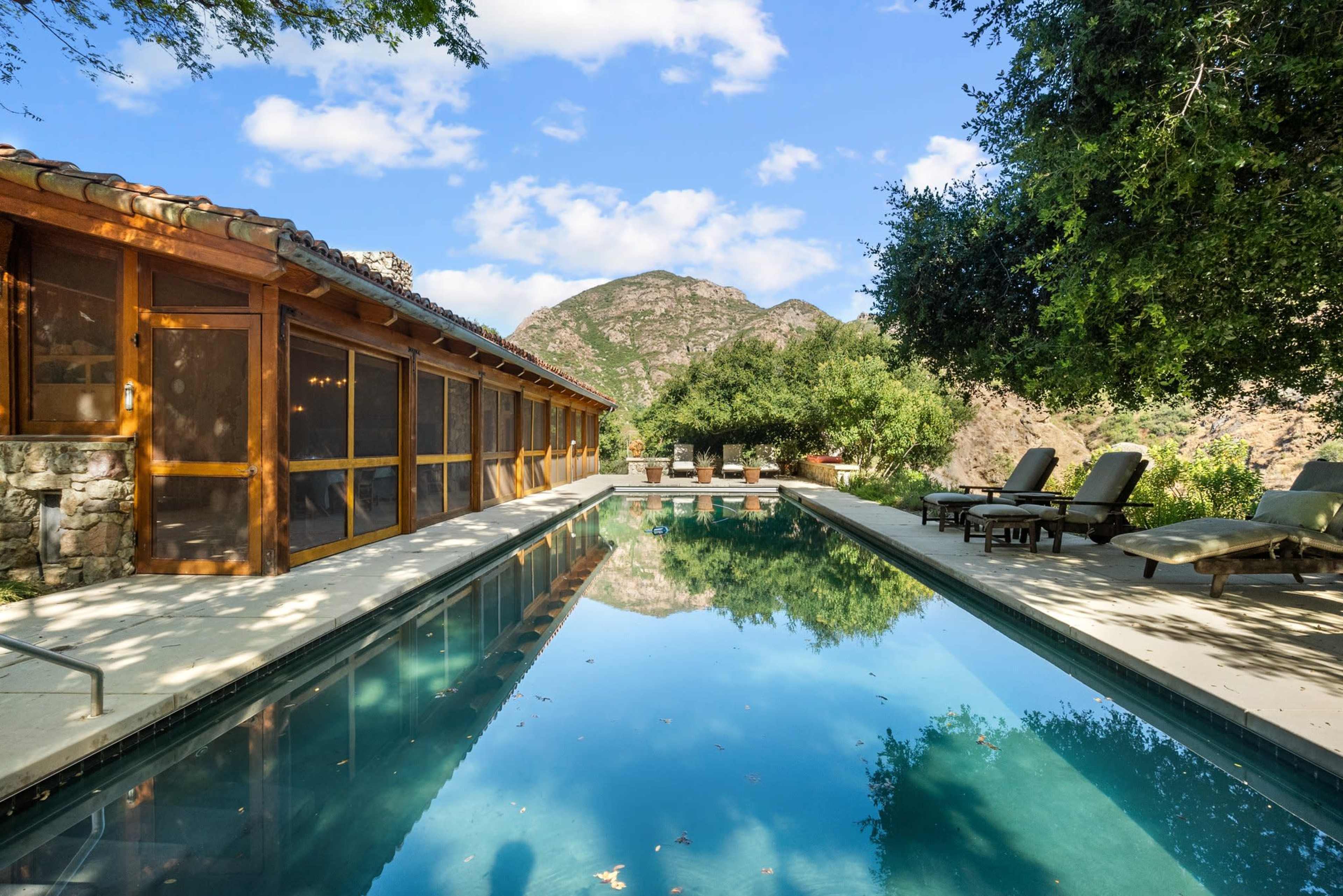 The image shows a tranquil outdoor area featuring a swimming pool, surrounded by a wooden structure, lounge chairs, and lush greenery, with mountains in the background.