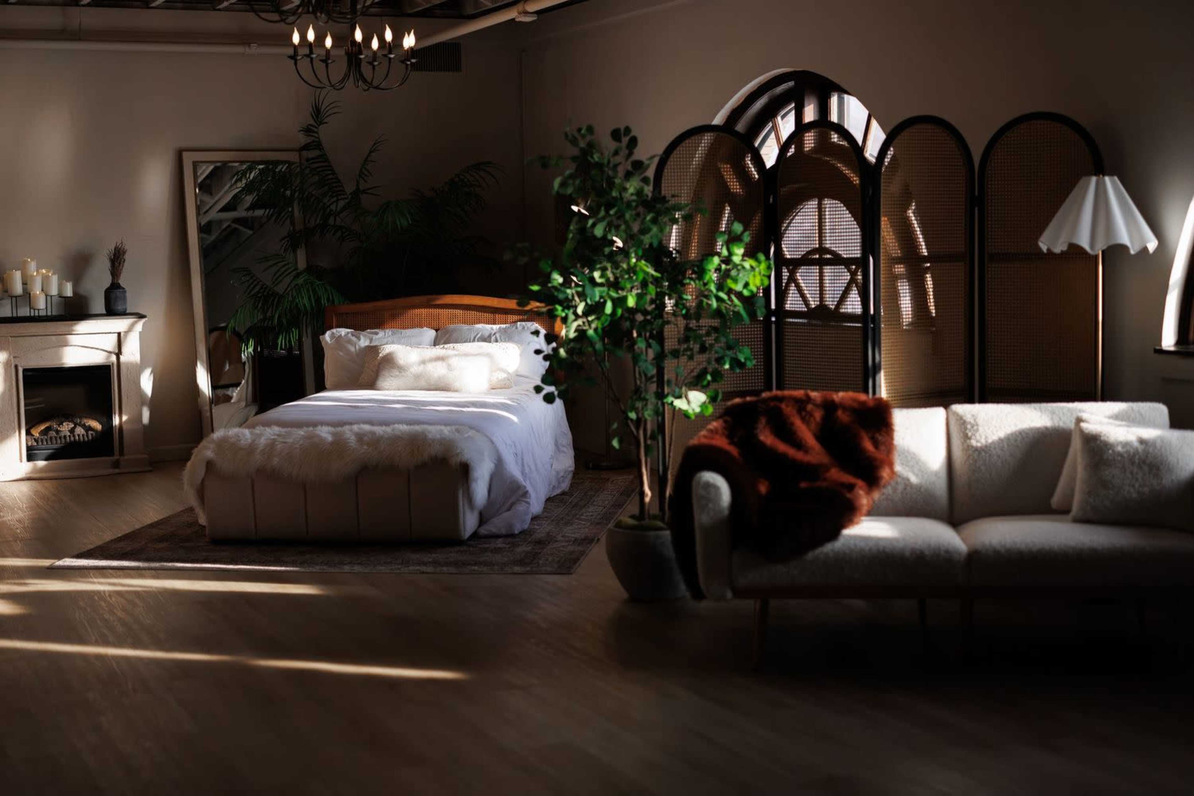 The image shows a softly lit bedroom featuring a bed with white bedding, a green plant, a cozy sofa, and a decorative screen in a spacious, stylishly designed room.
