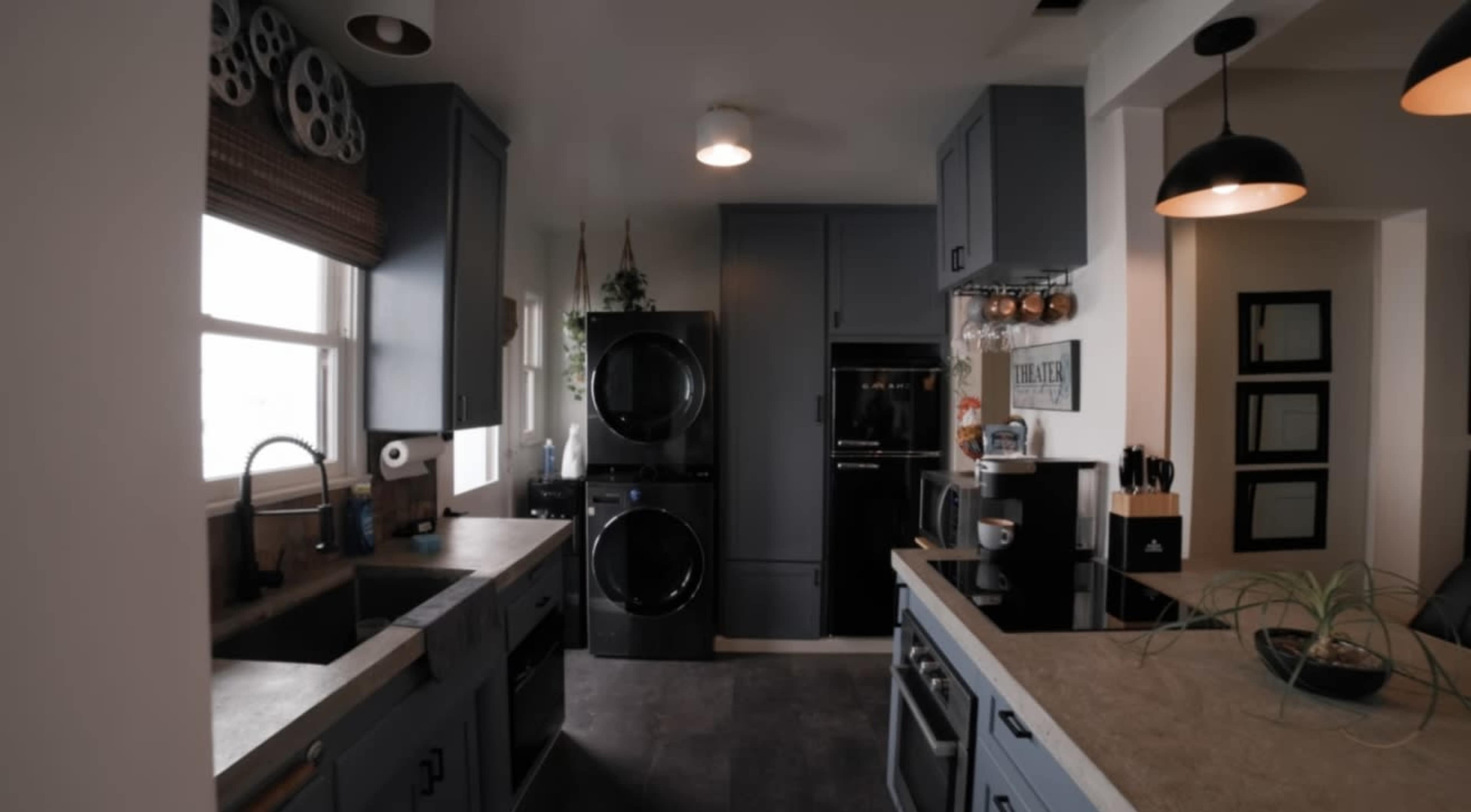 The image shows a modern kitchen with gray cabinetry, a double sink, and a laundry area featuring stacked black appliances.