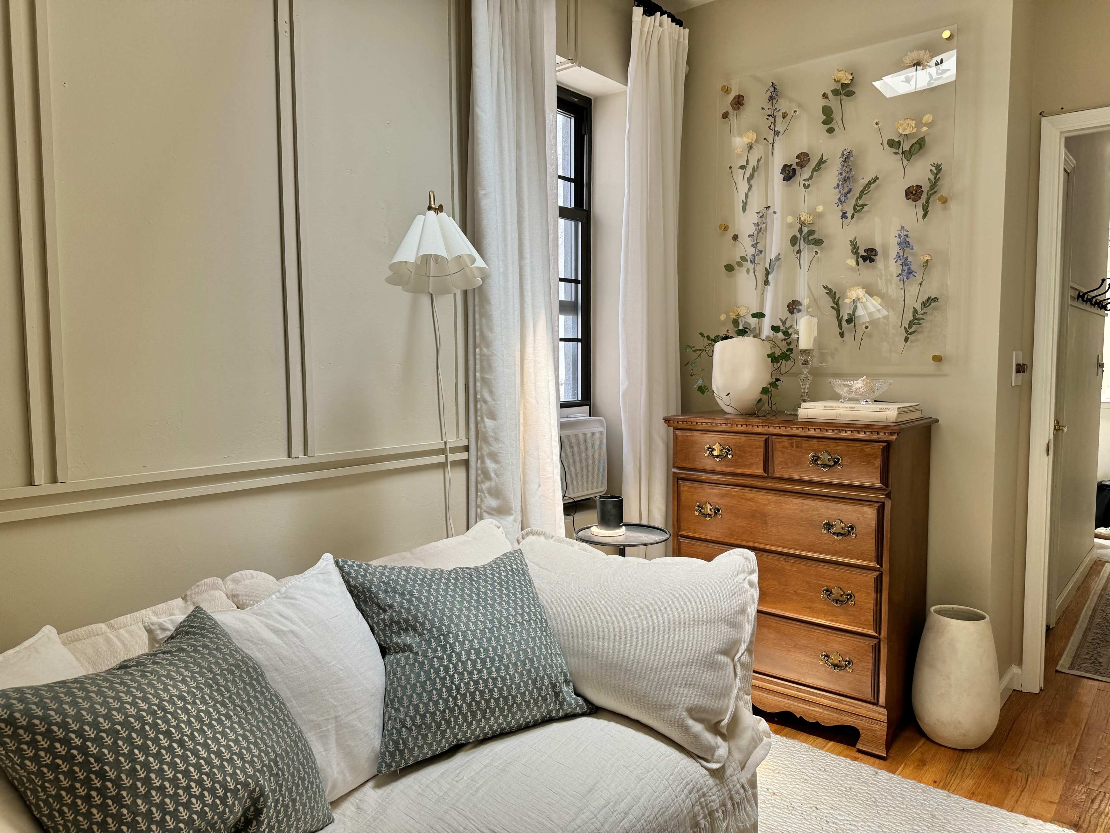 The scene features a sunlit room with a light-colored sofa, decorative pillows, a wooden dresser, and a wall adorned with floral decorations.