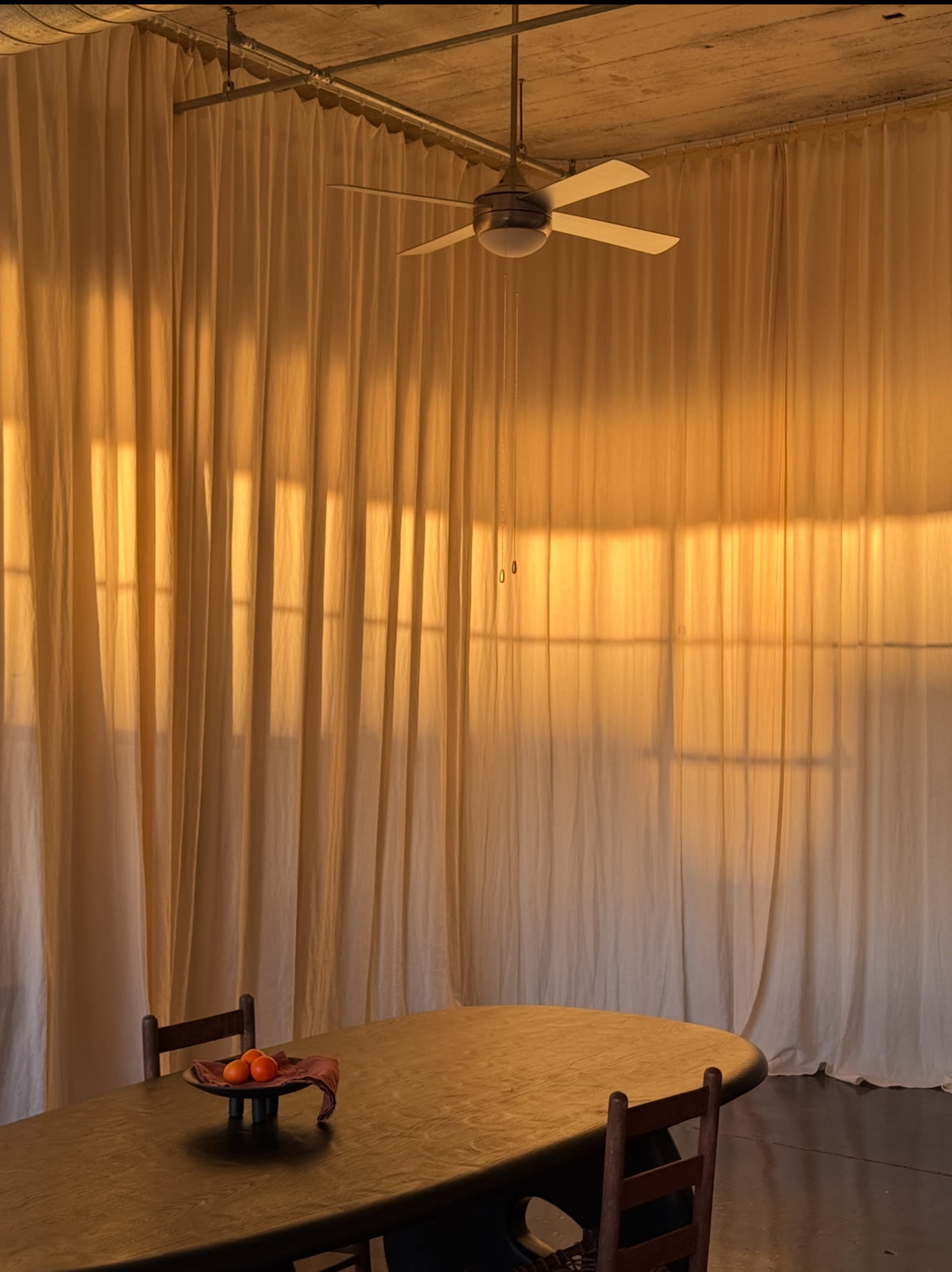 A round table with a bowl of oranges sits in a softly lit room with sheer curtains casting shadows on the wall.