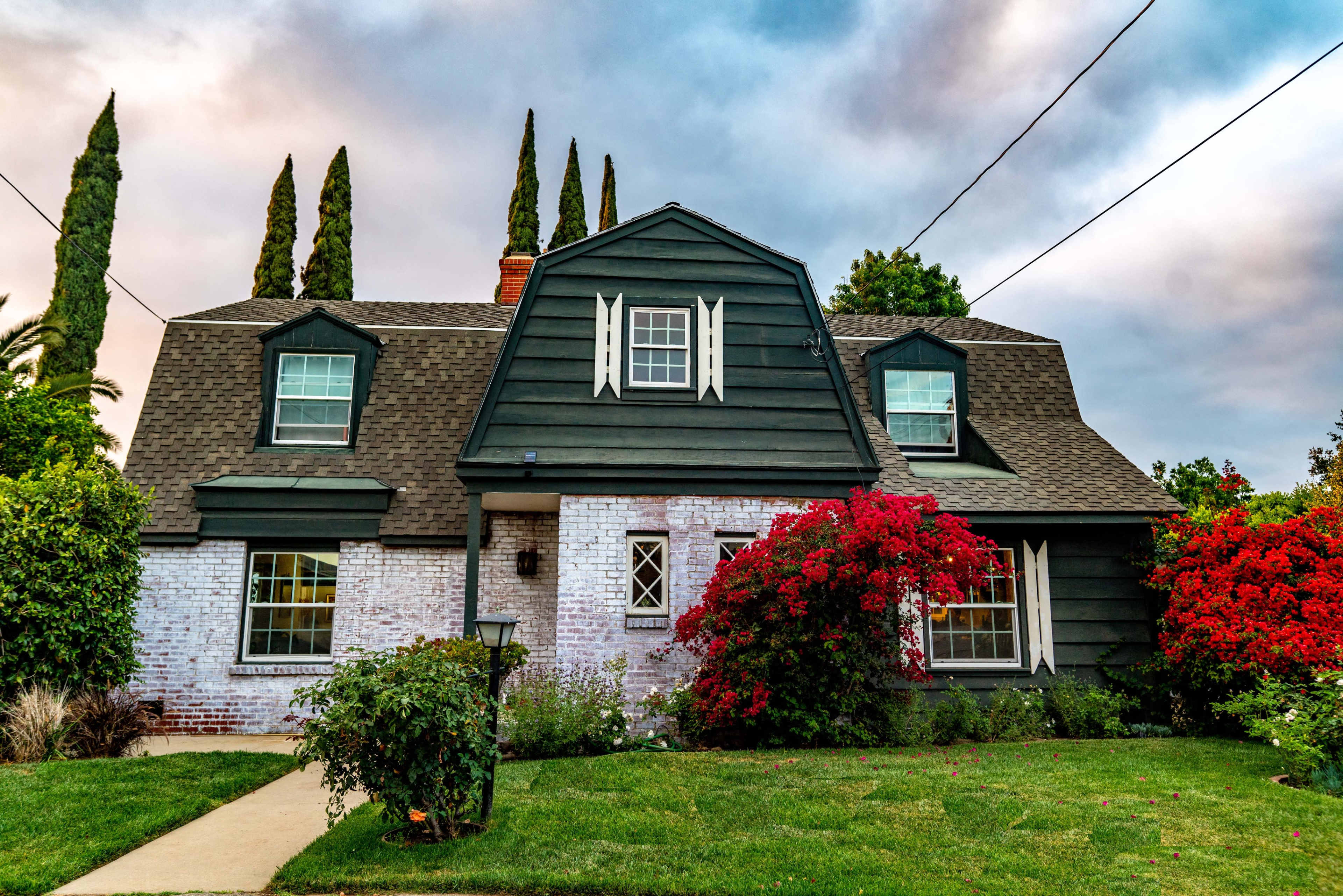 A two-story house with green siding, a gray roof, and red flowering bushes in the front yard.