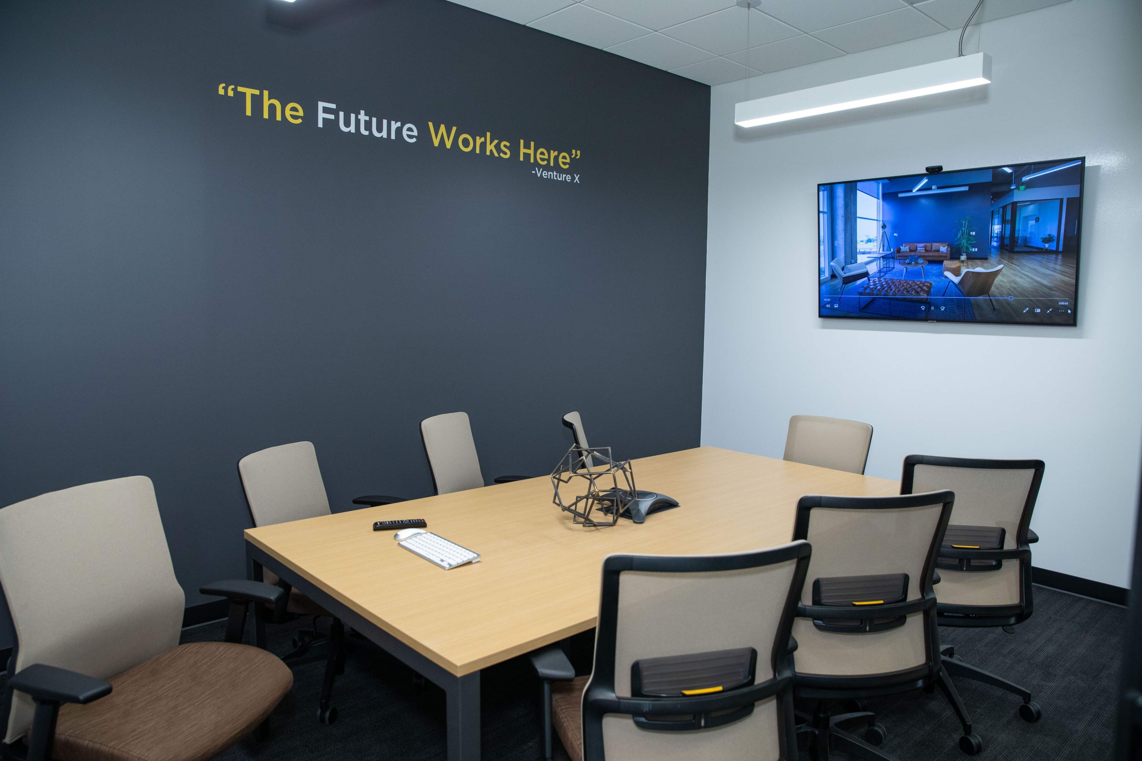 A conference room features a table surrounded by chairs, with a large screen displaying an image on the wall next to a quote about the future.