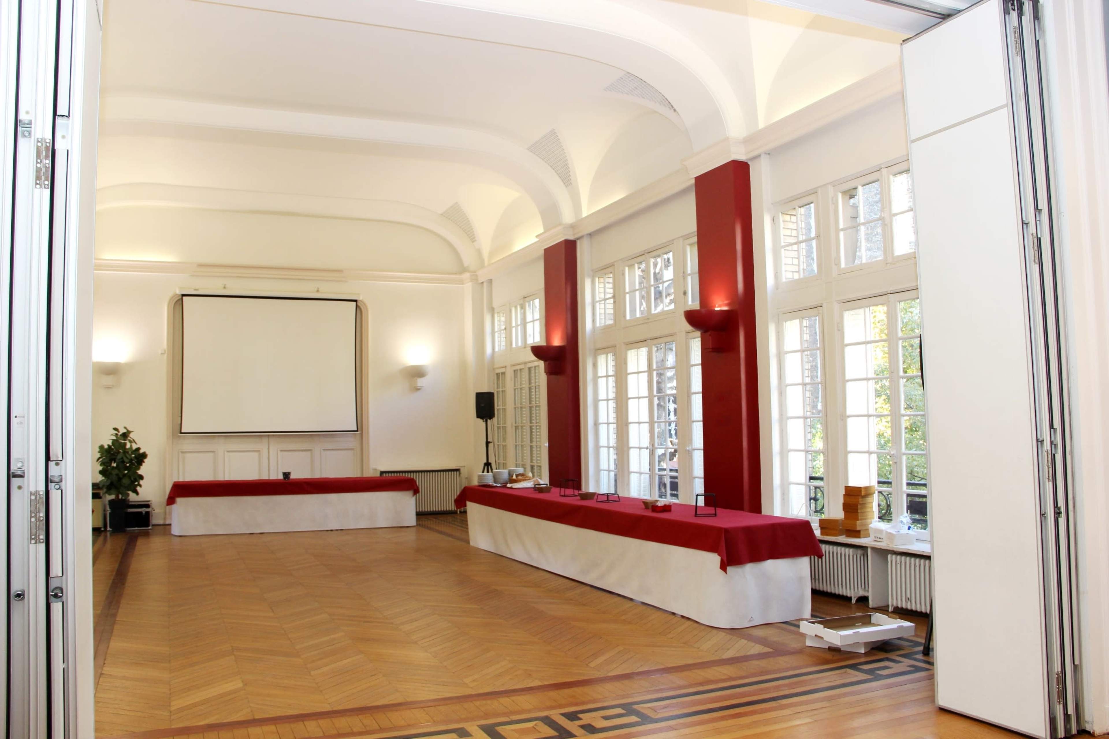 A spacious room features a large screen and a long table covered with a red cloth, flanked by large windows letting in natural light.
