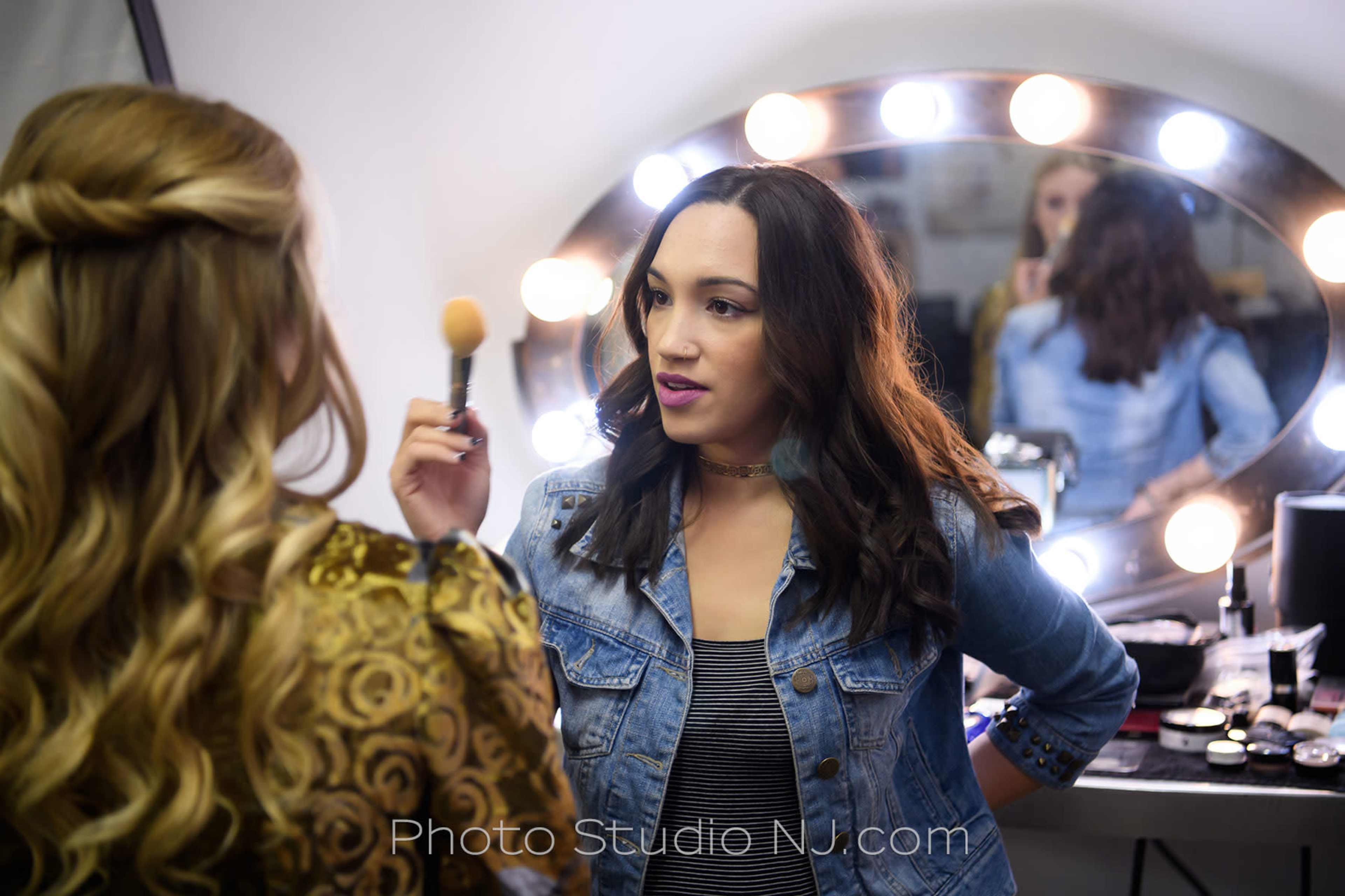 A makeup artist applies makeup to a client in front of a well-lit vanity mirror.