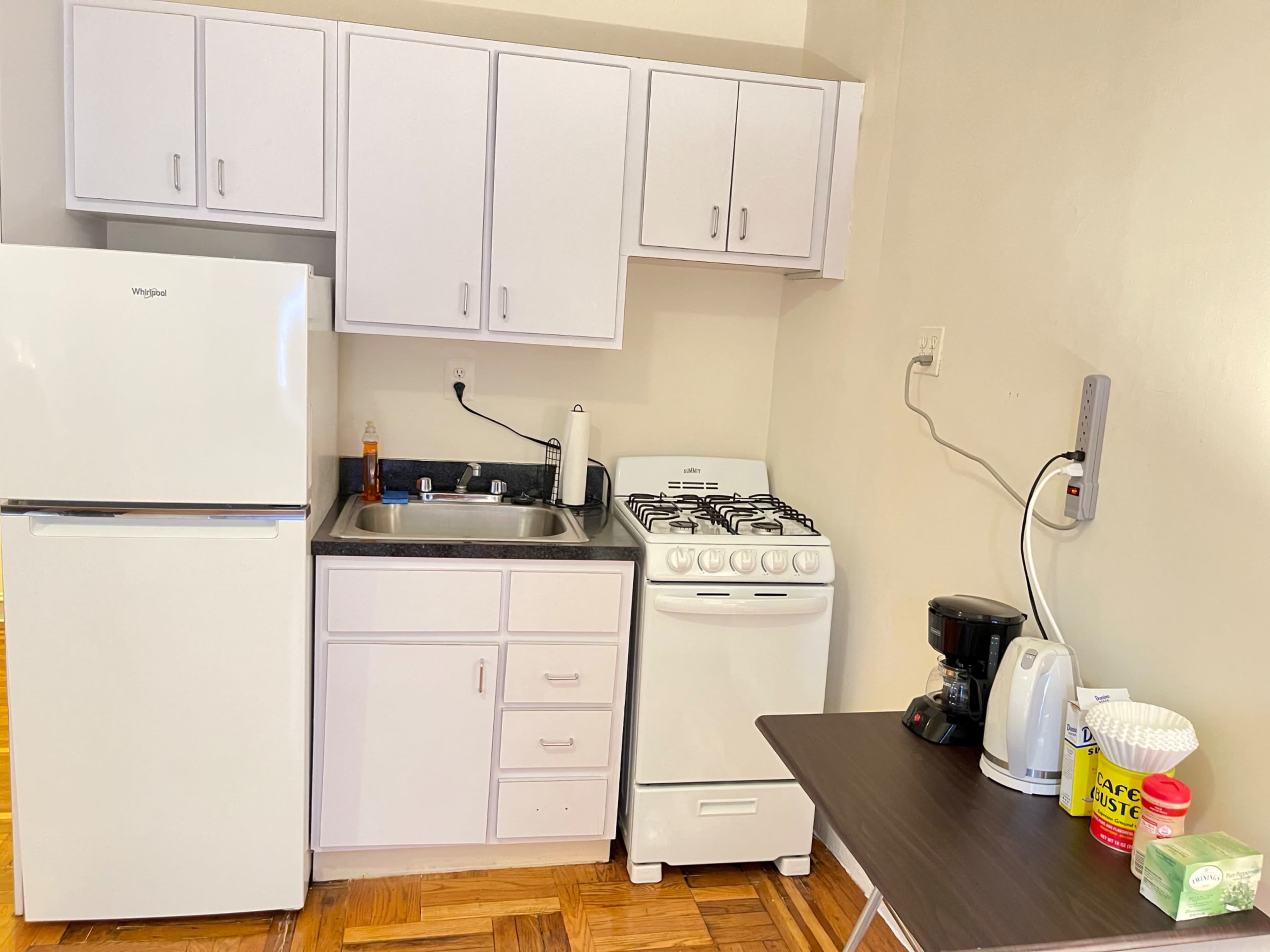 The image shows a compact kitchen with a white refrigerator, sink, gas stove, coffee maker, and a small table.