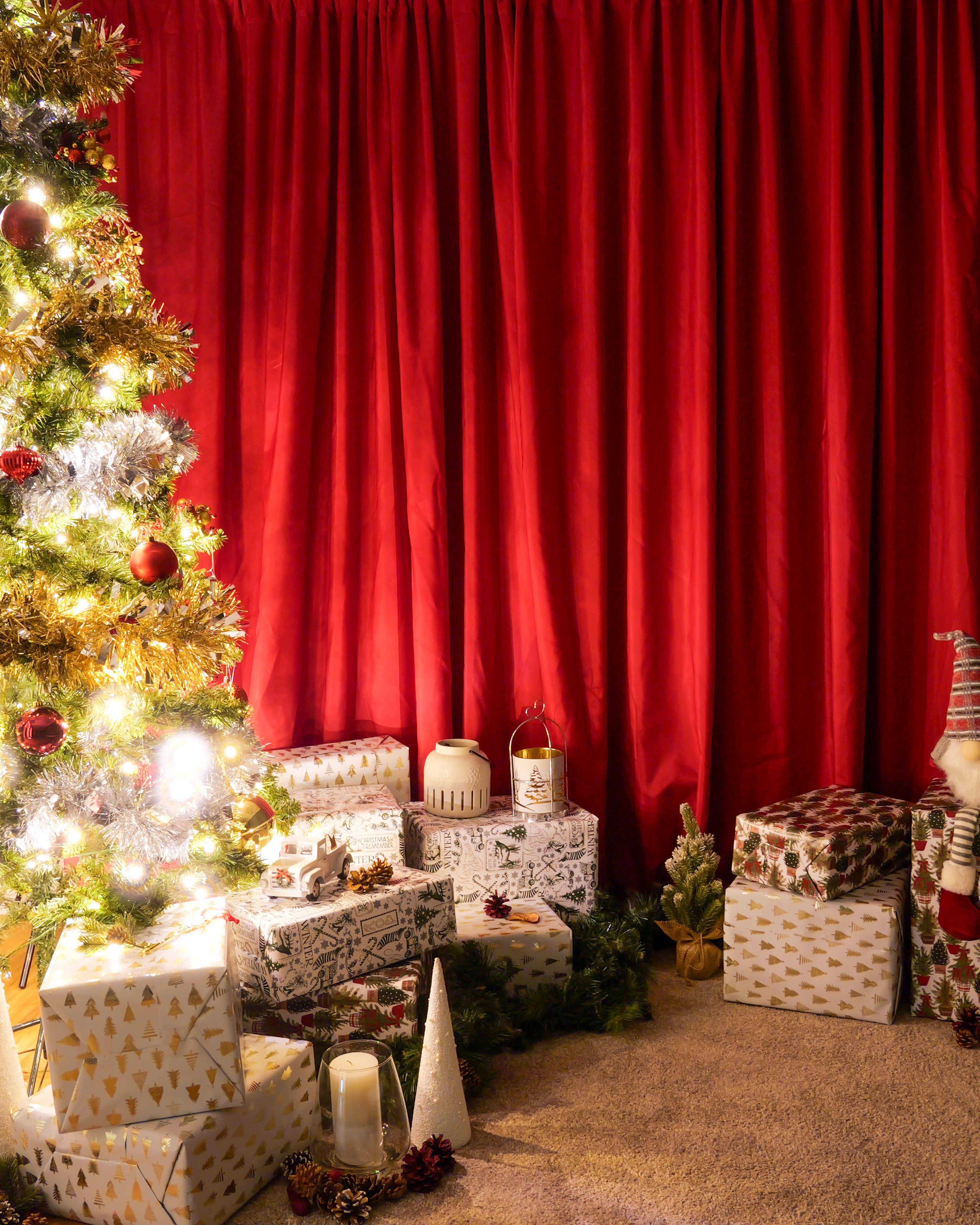 A decorated Christmas tree stands beside neatly wrapped gifts on the floor, with a backdrop of rich red curtains.