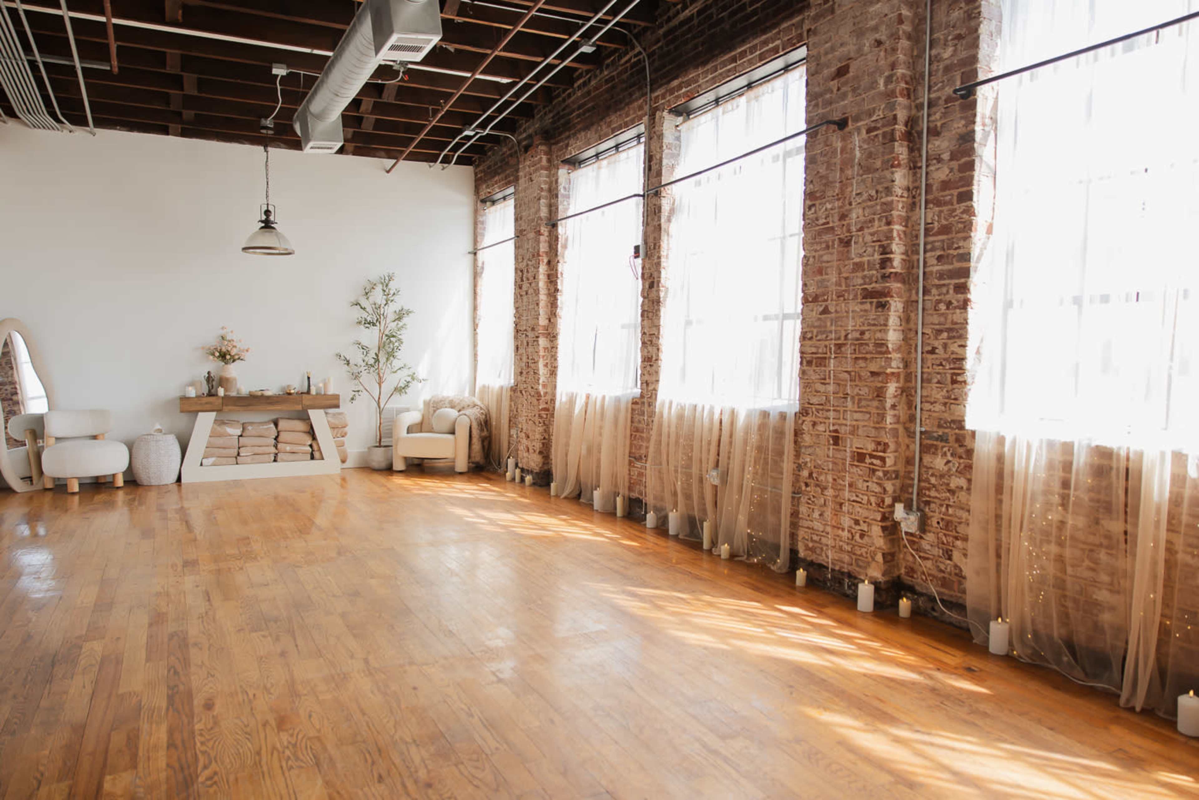 The image shows a spacious, well-lit interior with wooden floors, large windows adorned with sheer curtains, and minimal furniture, including a sofa and a decorative table.