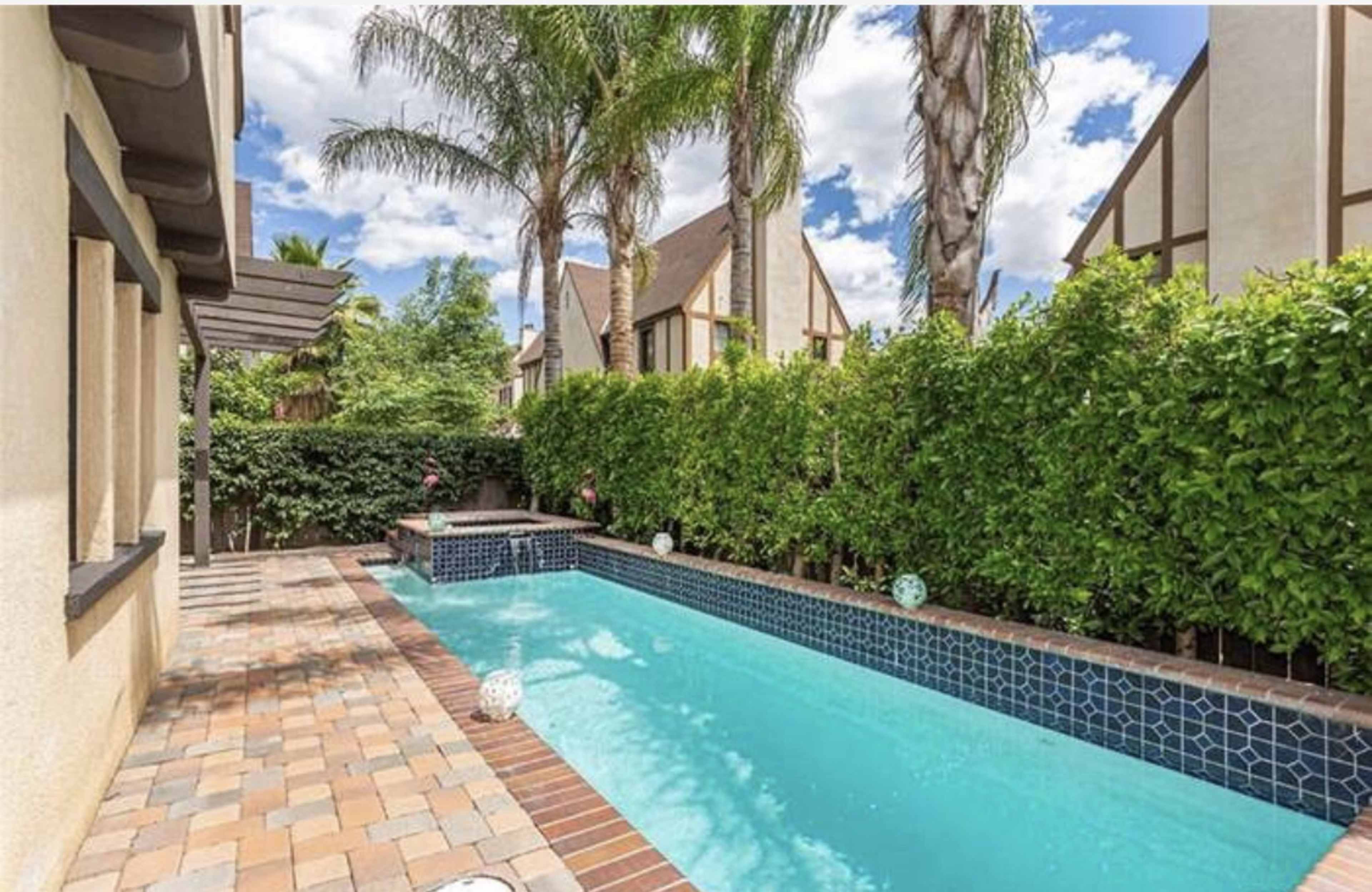 A backyard swimming pool surrounded by lush greenery and palm trees, with a brick patio in the foreground.