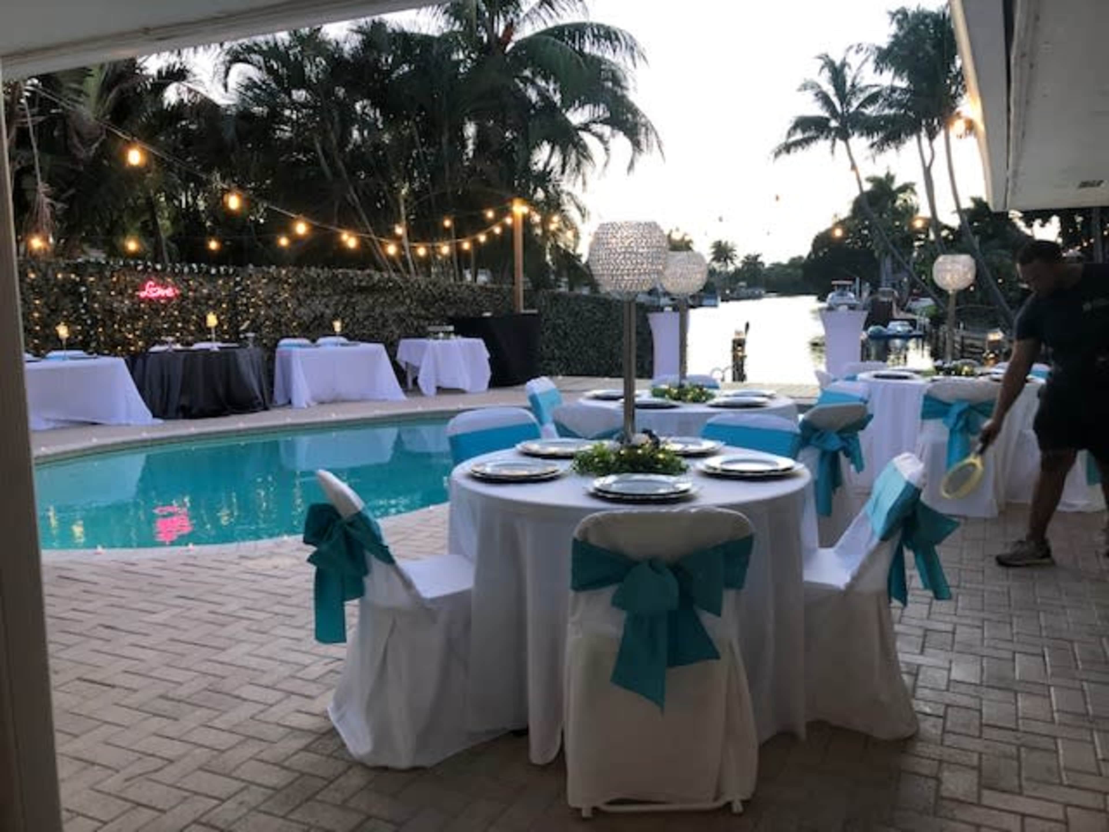 A poolside event space is set up with tables draped in white linens and adorned with turquoise ribbons, surrounded by tropical plants and string lights.