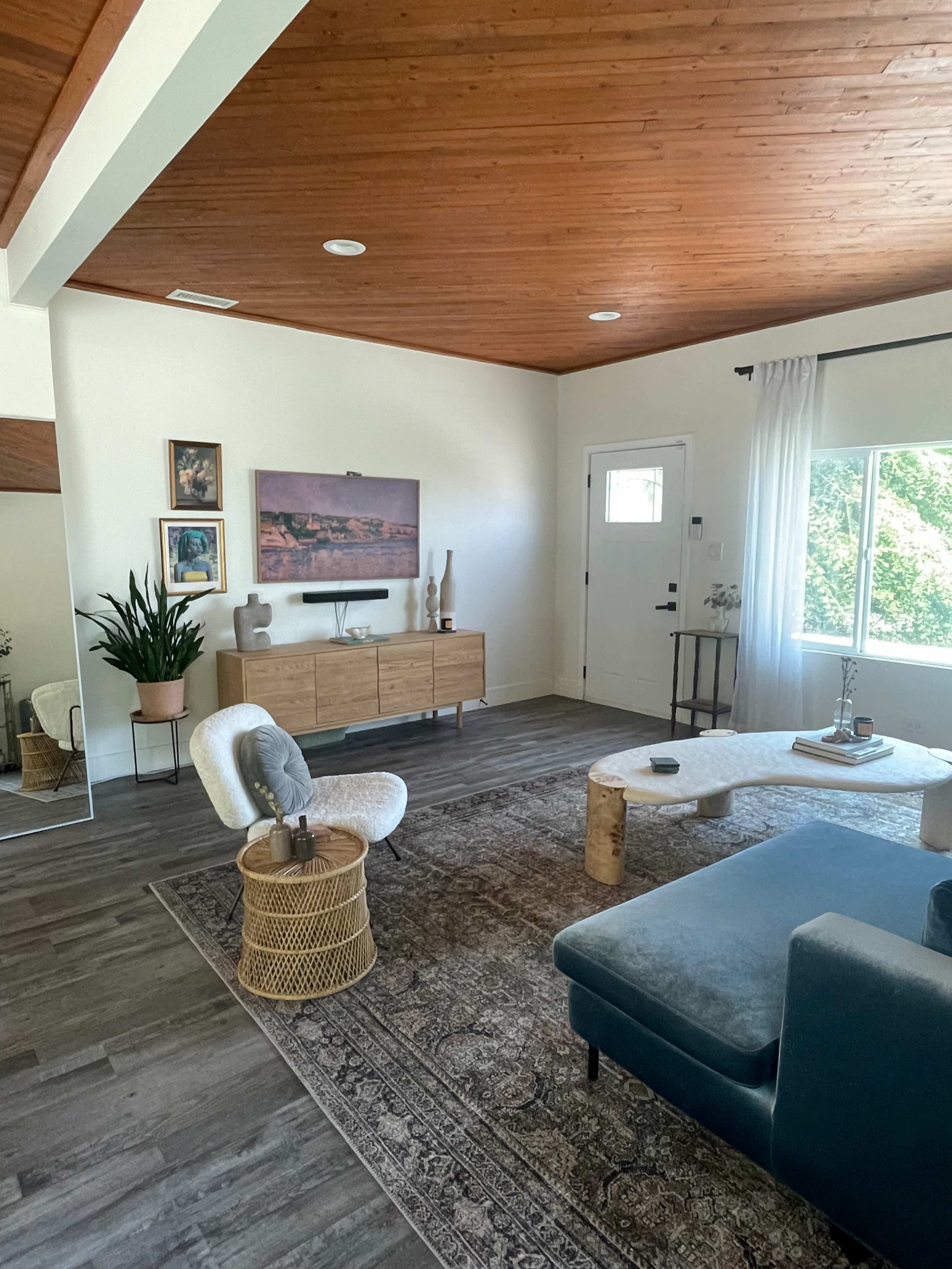 The image shows a modern living room with a wooden ceiling, a large window, a green sofa, a light-colored coffee table, and a tall planter with a leafy plant.