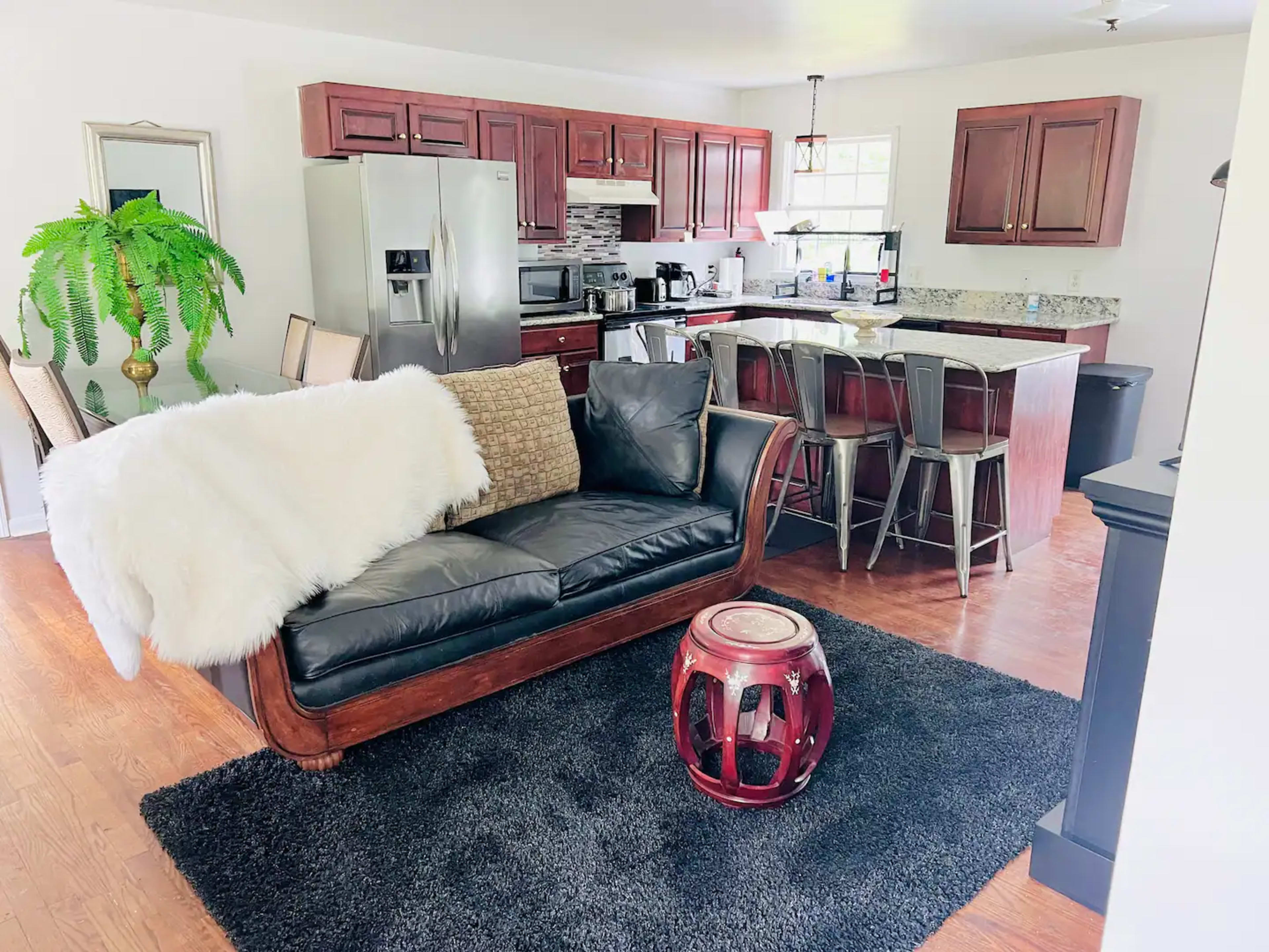 A modern open-plan living area with a black leather sofa, a woven throw, a round red table, and a kitchen area featuring wooden cabinets and stainless steel appliances.