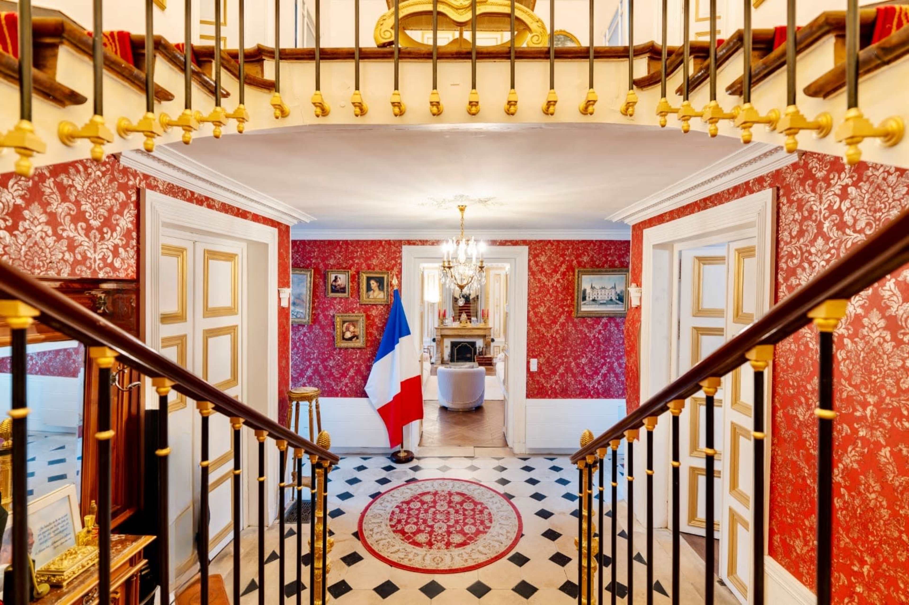 The image shows a lavishly decorated foyer with a grand staircase, ornate wallpaper, and a French flag positioned beside a colorful circular rug.