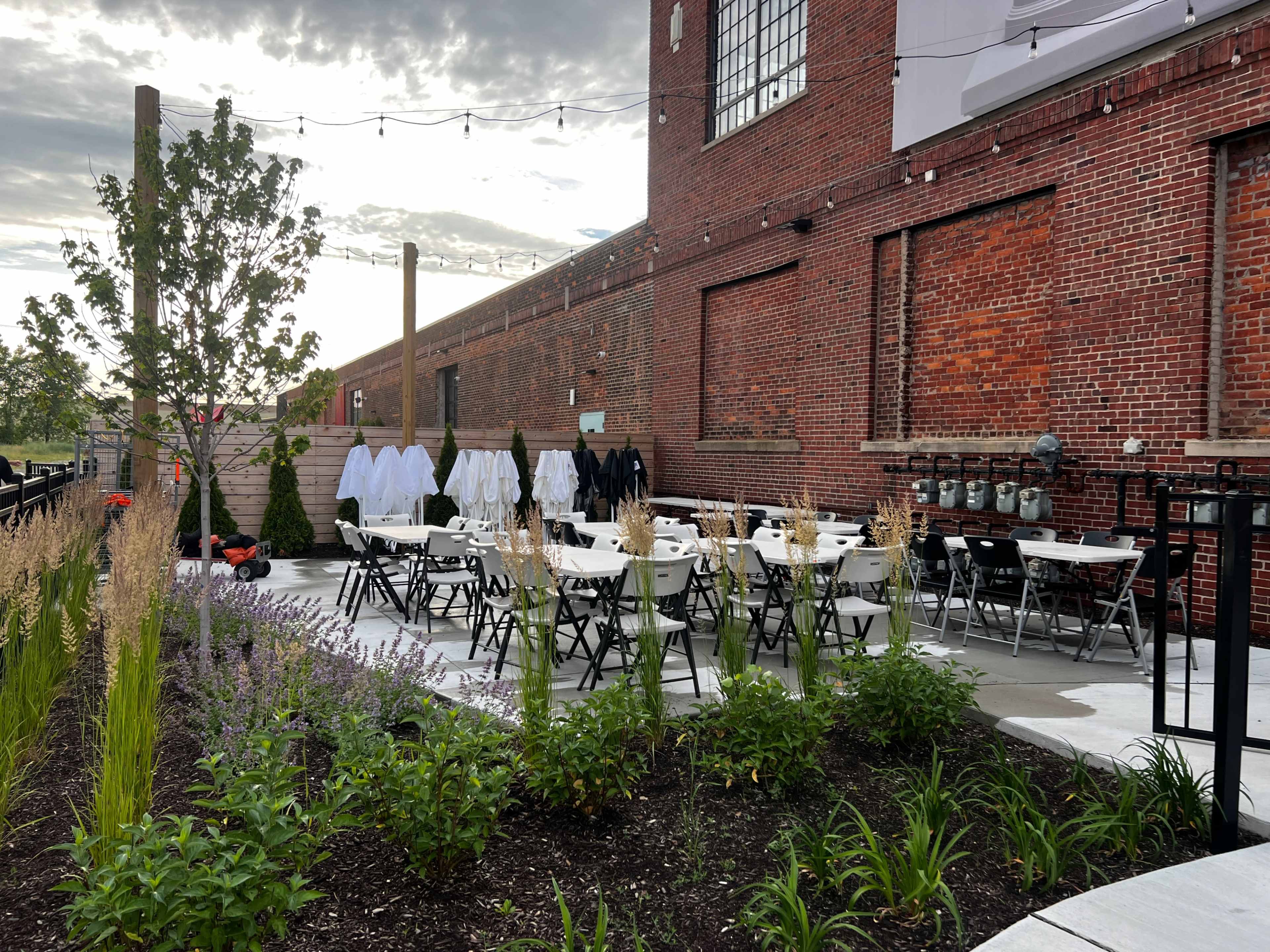 An outdoor dining area is set up with tables and chairs, surrounded by plants and a brick building in the background.