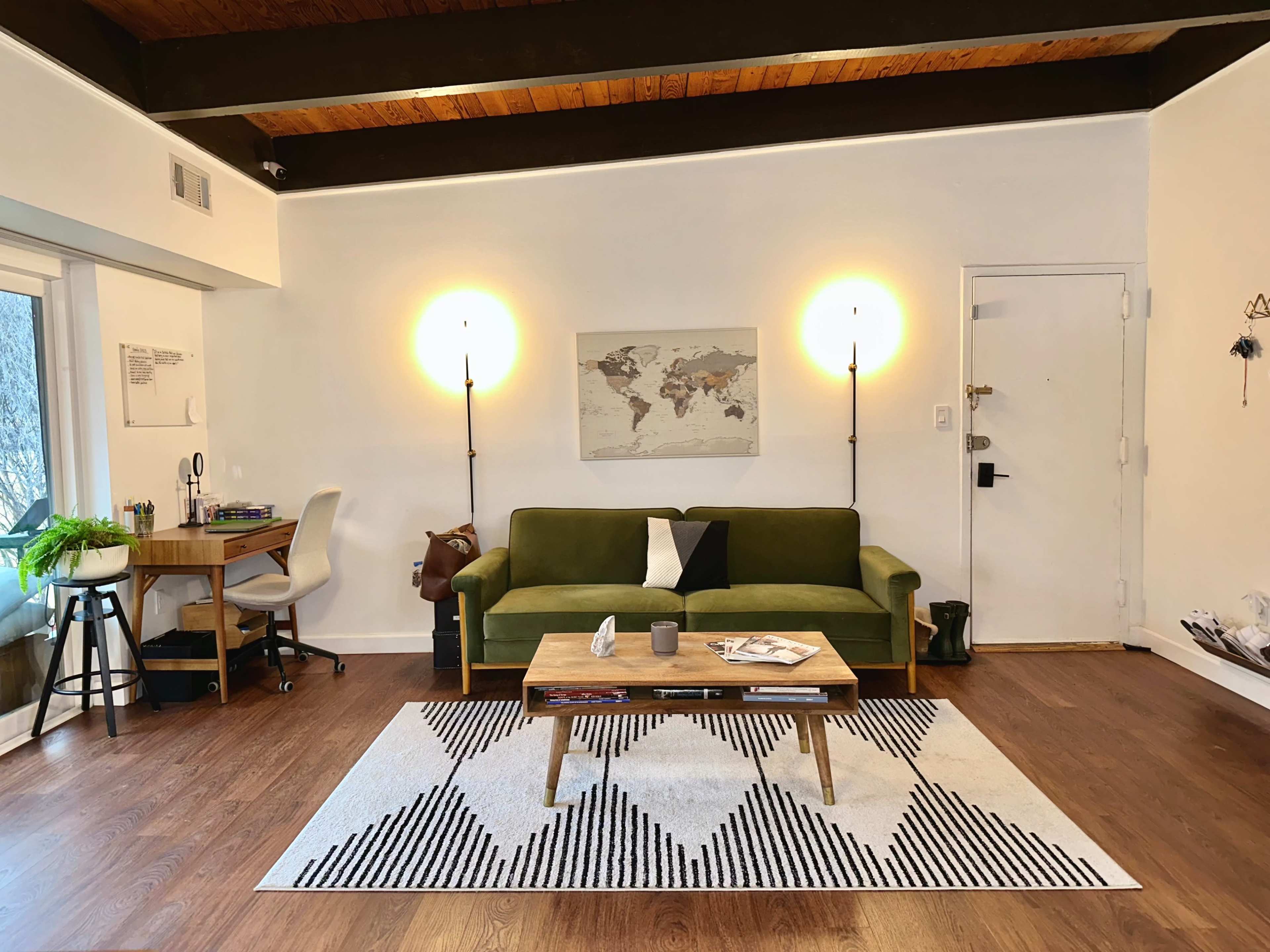 A living room with a green sofa, a wooden coffee table with decorative items, and a desk in the corner beside a large window.
