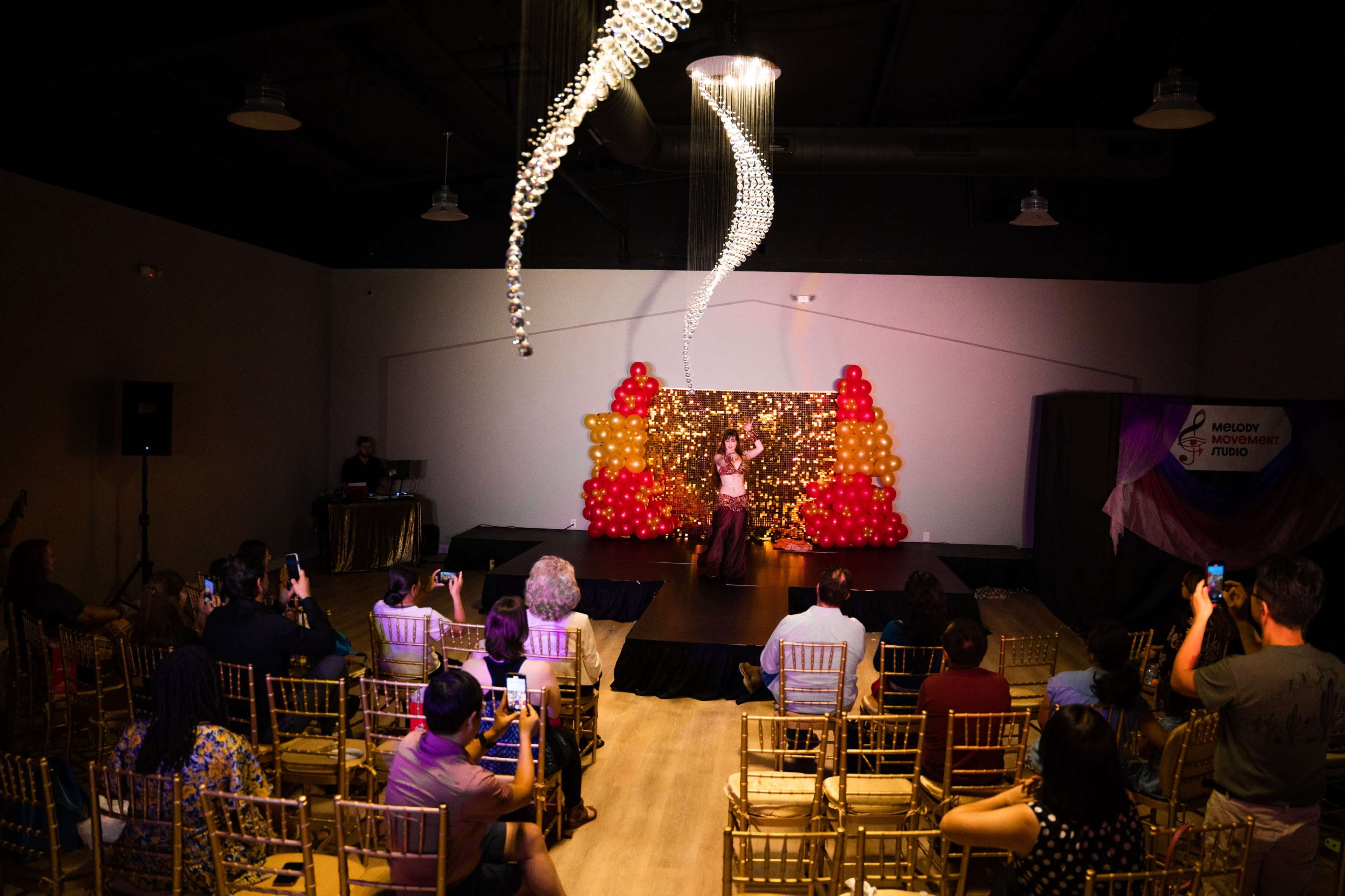A performer stands on a stage in front of a colorful balloon backdrop while an audience captures the moment with their phones.