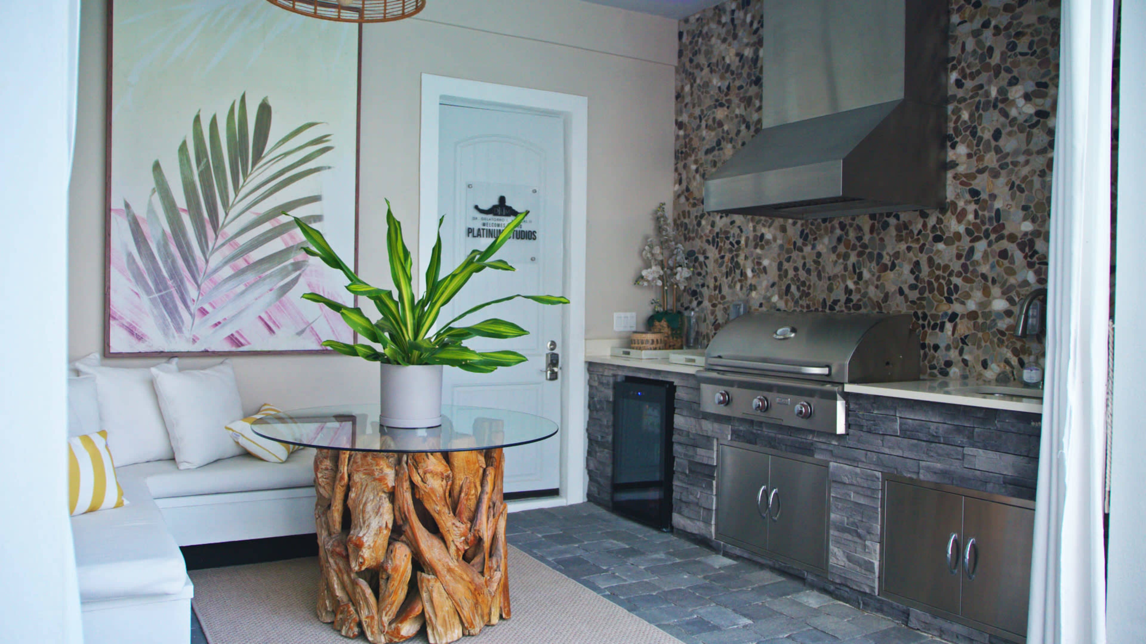 The image shows an outdoor kitchen area featuring a stainless steel grill, a stone accent wall, a circular glass table with a wooden base, and a potted plant, all arranged next to a seating area.