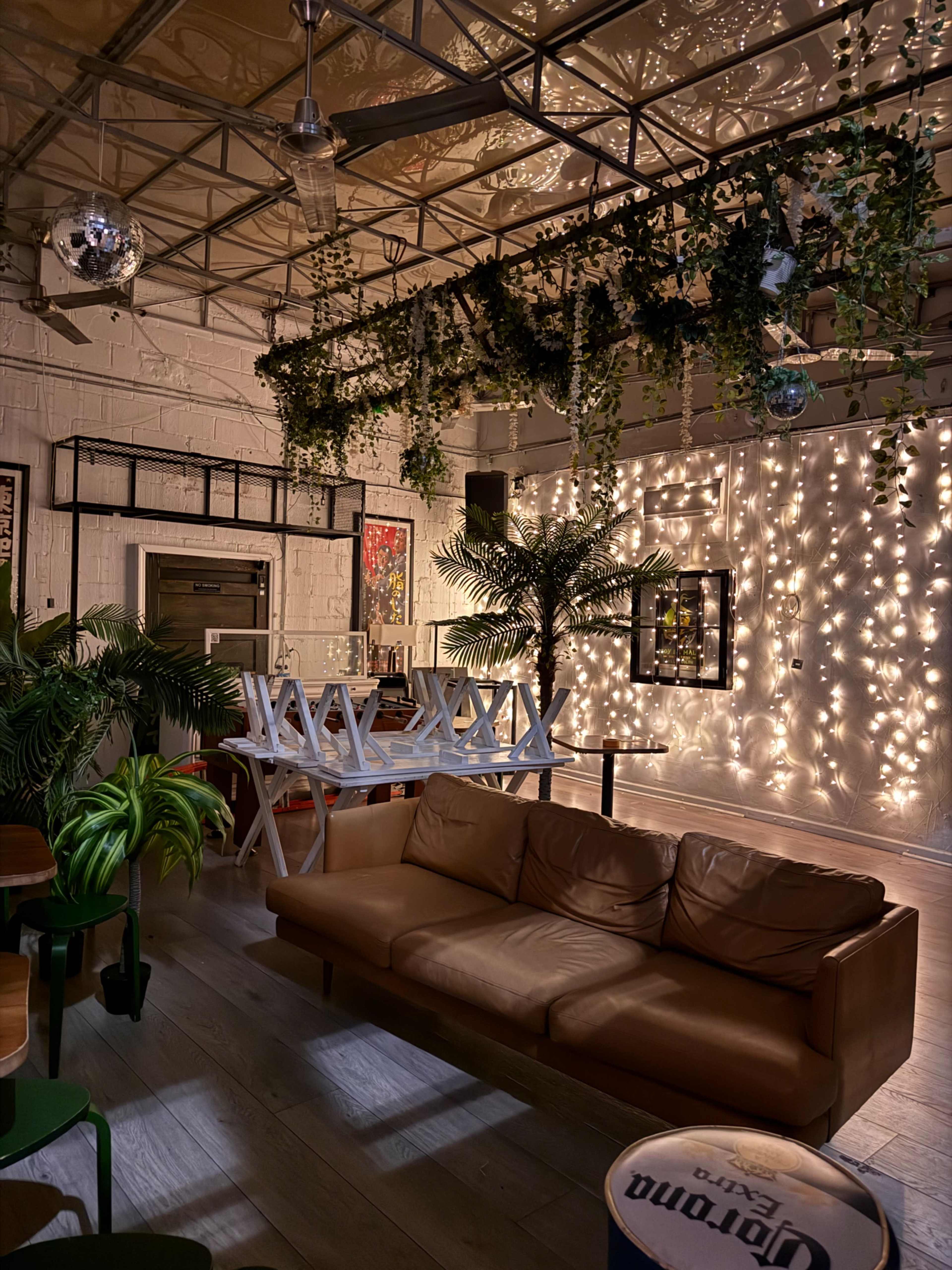 The image depicts a cozy indoor space with a brown leather couch, a white dining table, potted plants, and a wall adorned with string lights.